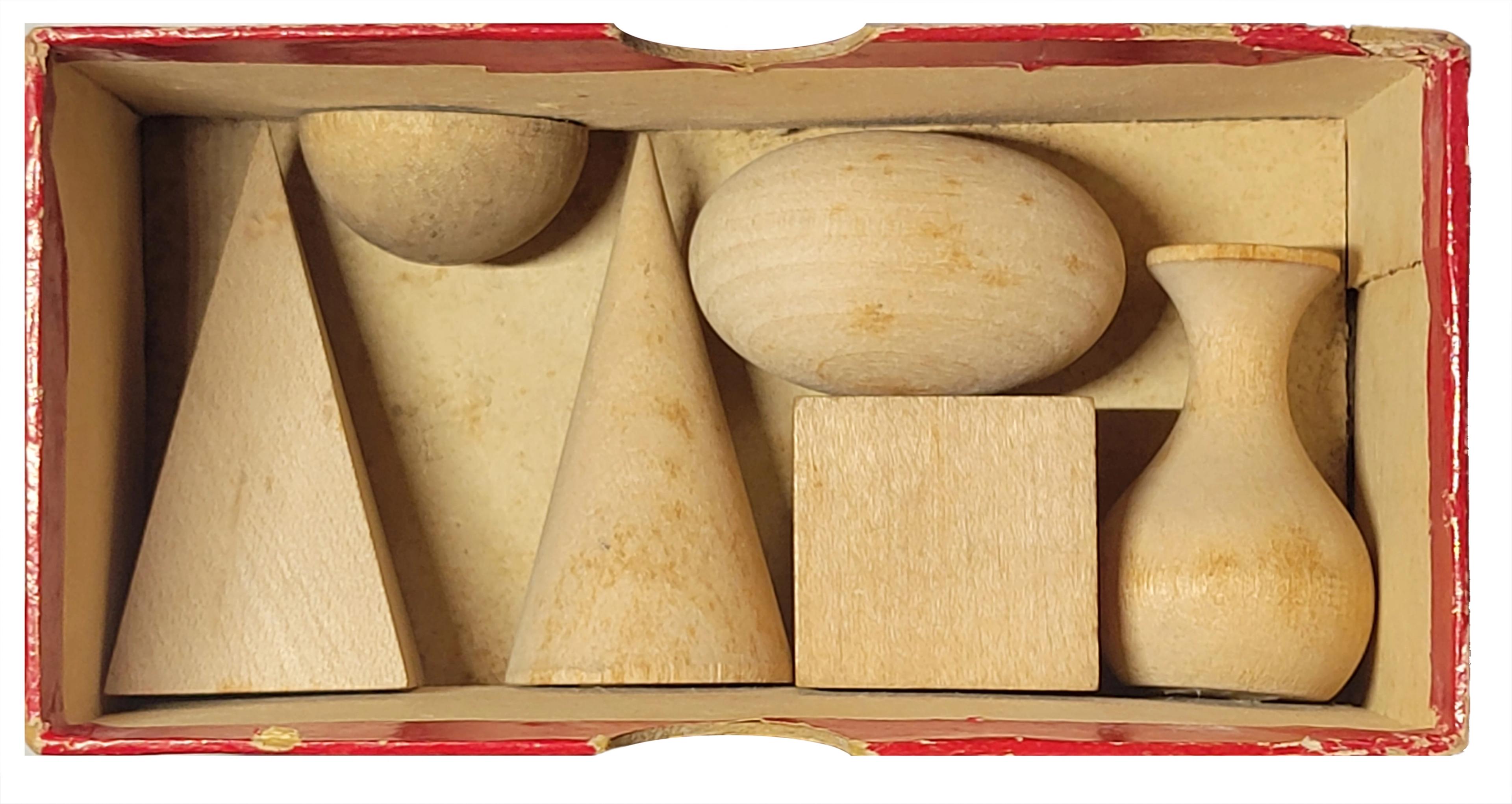 Unfinished wooden geometric forms, including cones, spheres, a cube, and a vase shape, fitted together inside a red-edged box.