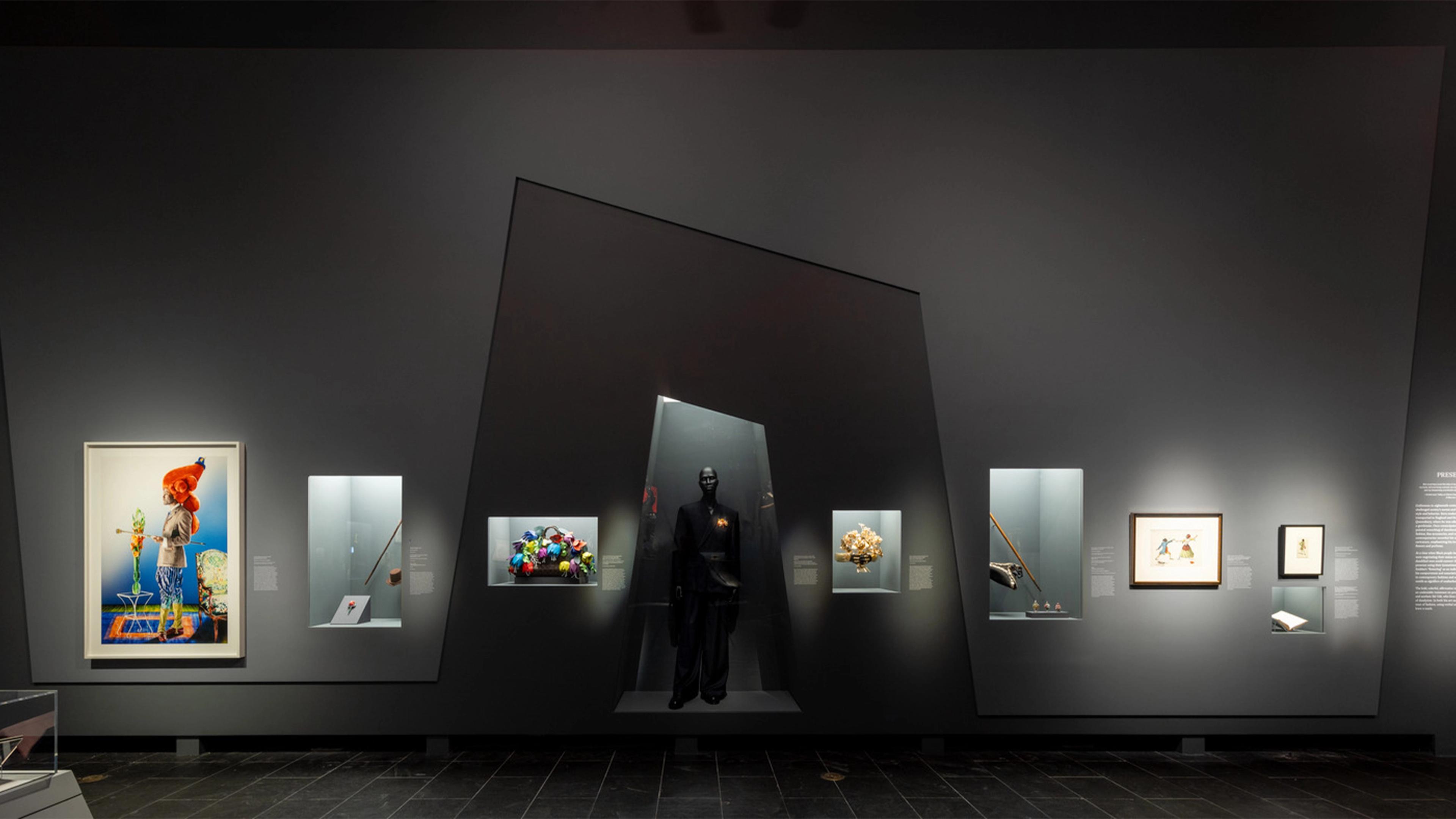 Exhibit with diverse artifacts on a dark wall: a colorful portrait, mannequin in black attire, various sculptures, and framed art, creating an enigmatic tone.