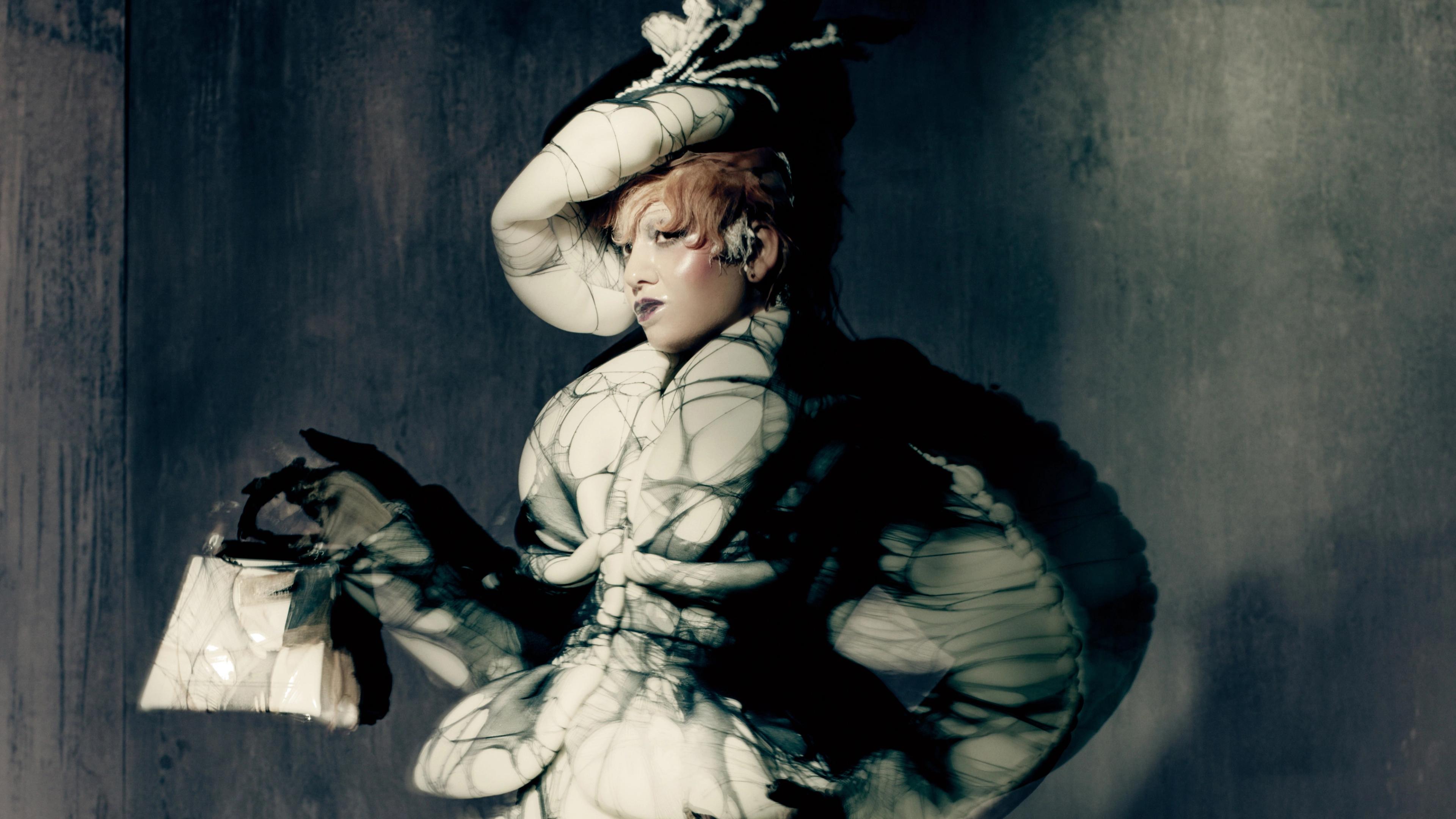 A model in a tailored suit jacket and hat with spider-weblike detailing holds a handbag,