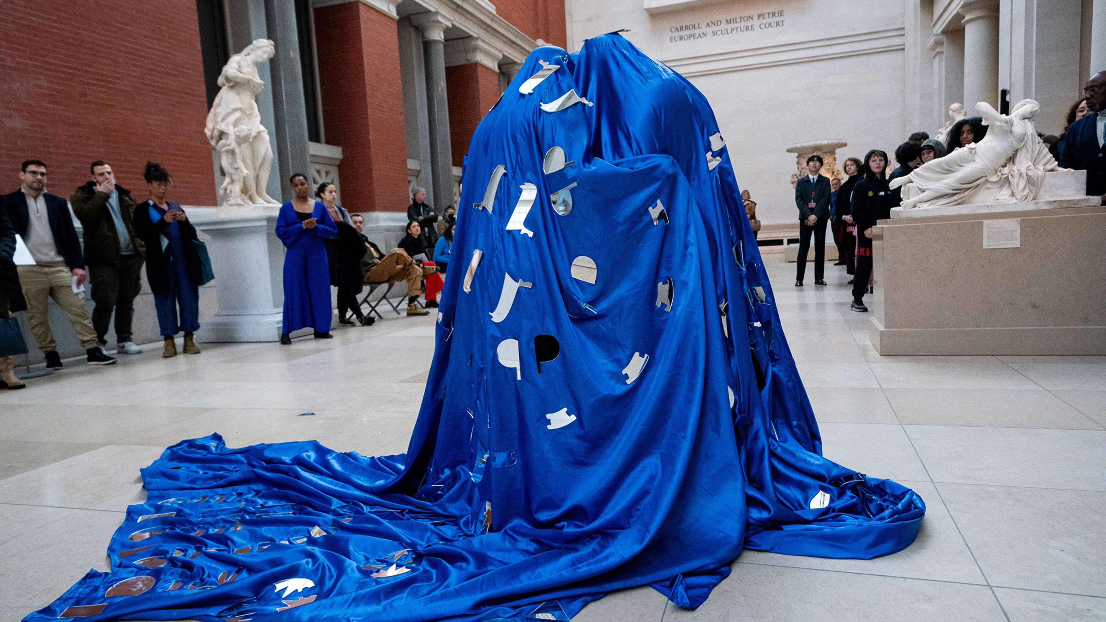 A figure shrouded in a royal blue, silky fabric with silver attachments in performance in the Petrie Sculpture Court