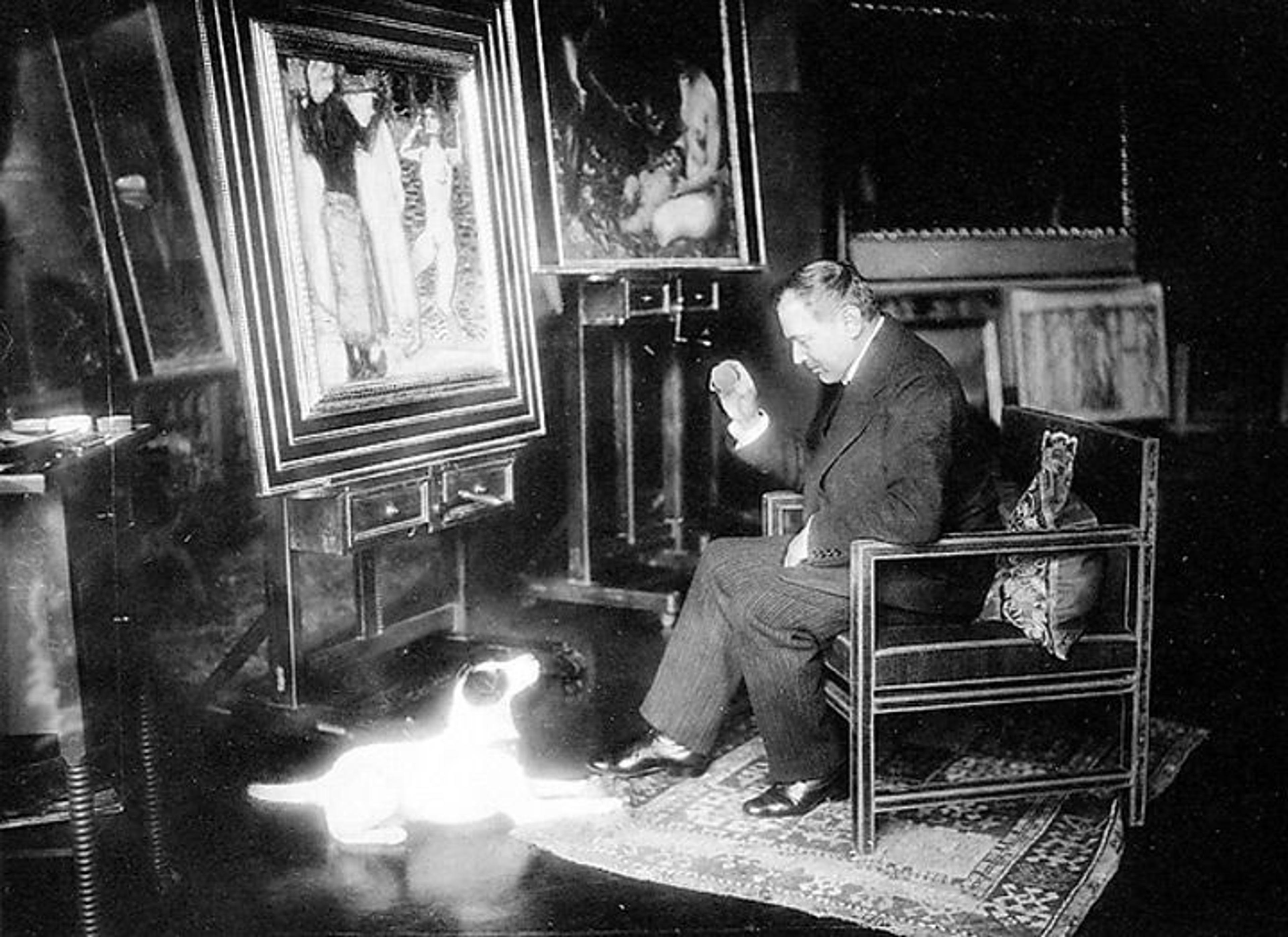Black-and-white archival photo of the artist Franz von Stuck sitting in a chair in his studio while looking at a small white dog on the floor