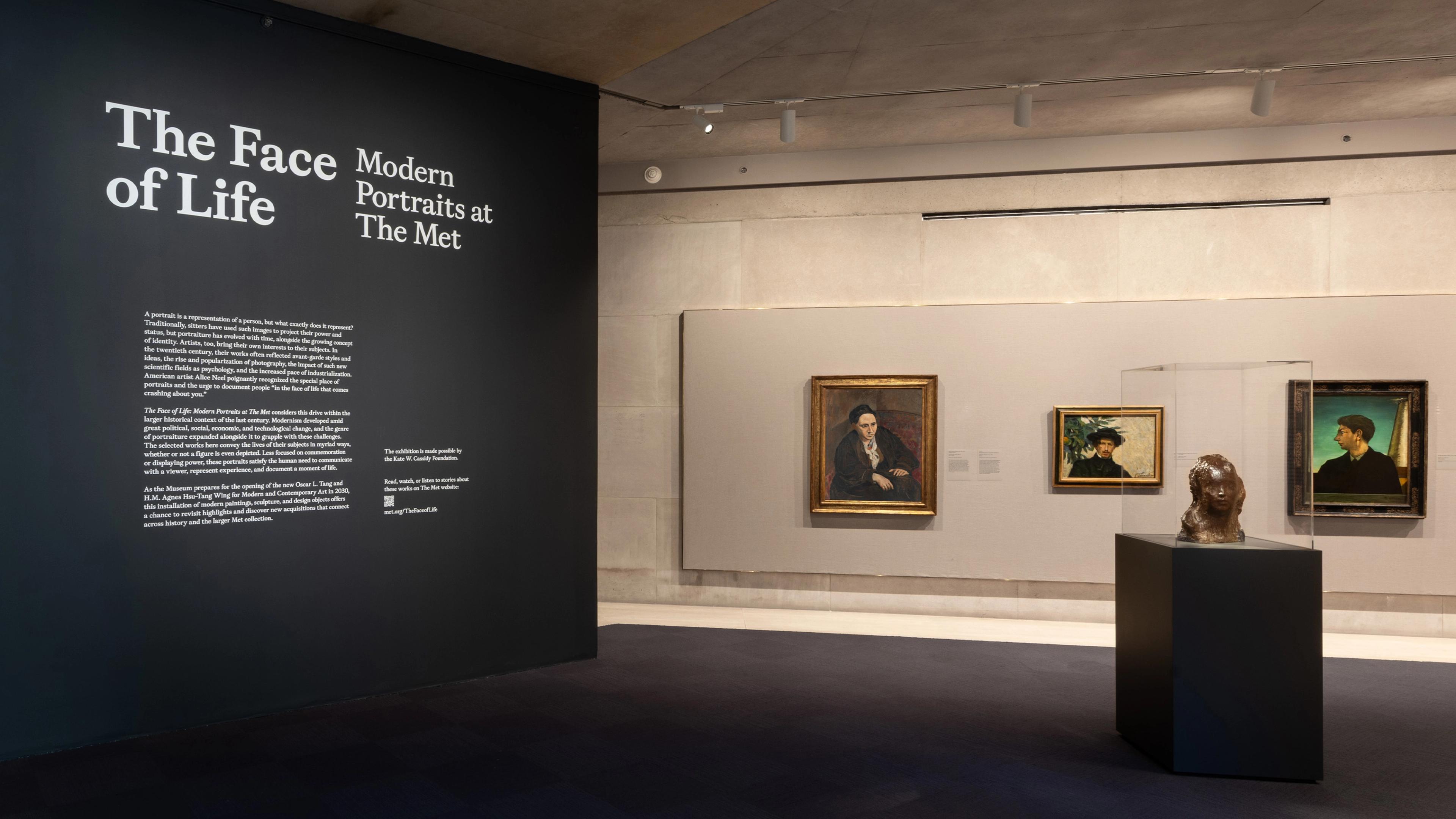 Exhibit features "The Face of Life: Modern Portraits at The Met." Dimly lit room with portraits on gray walls and a sculpture on a black pedestal.