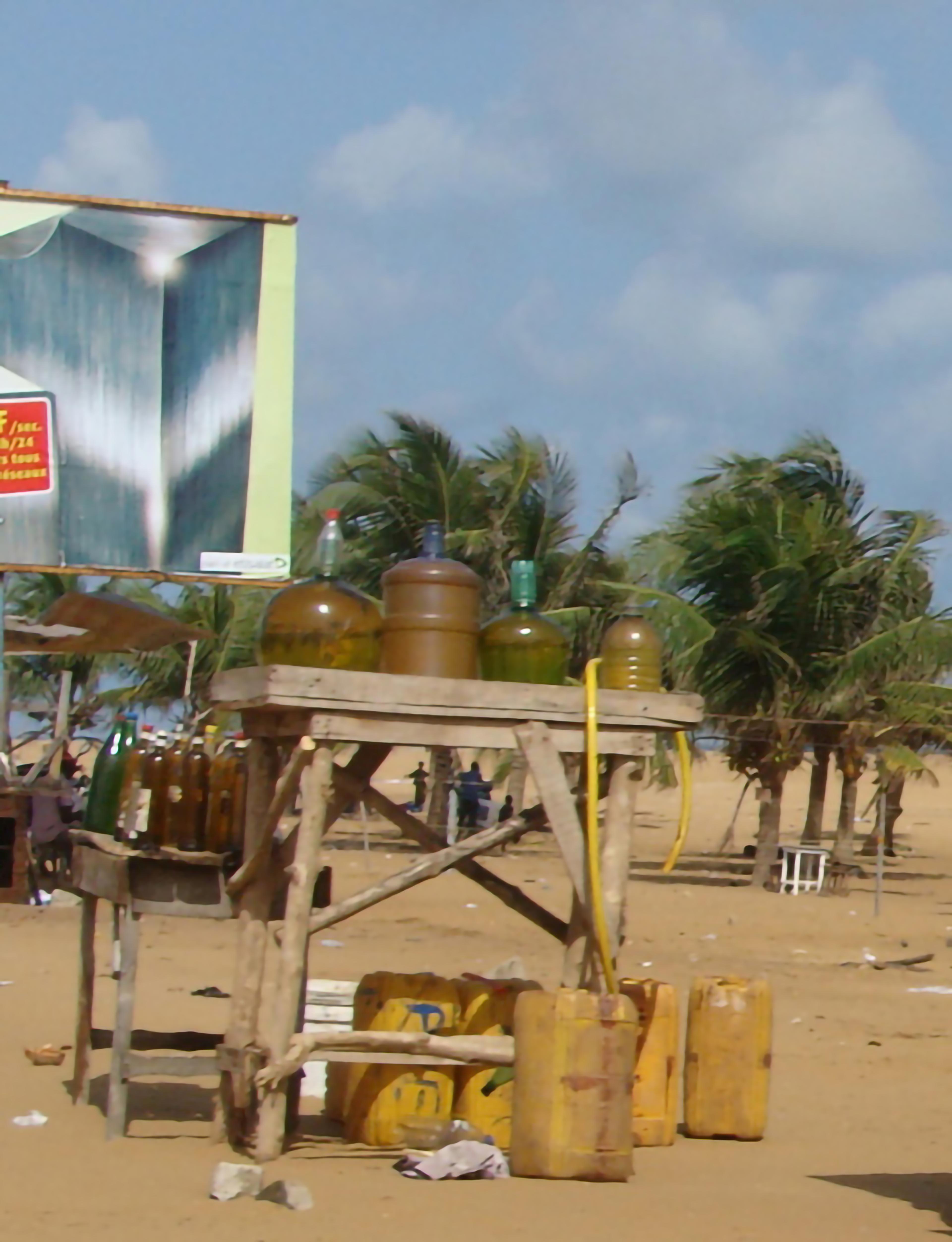 Two wooden tables with bottles and plastic jugs, set outdoors on sand. Palm trees and part of a billboard are in the background.