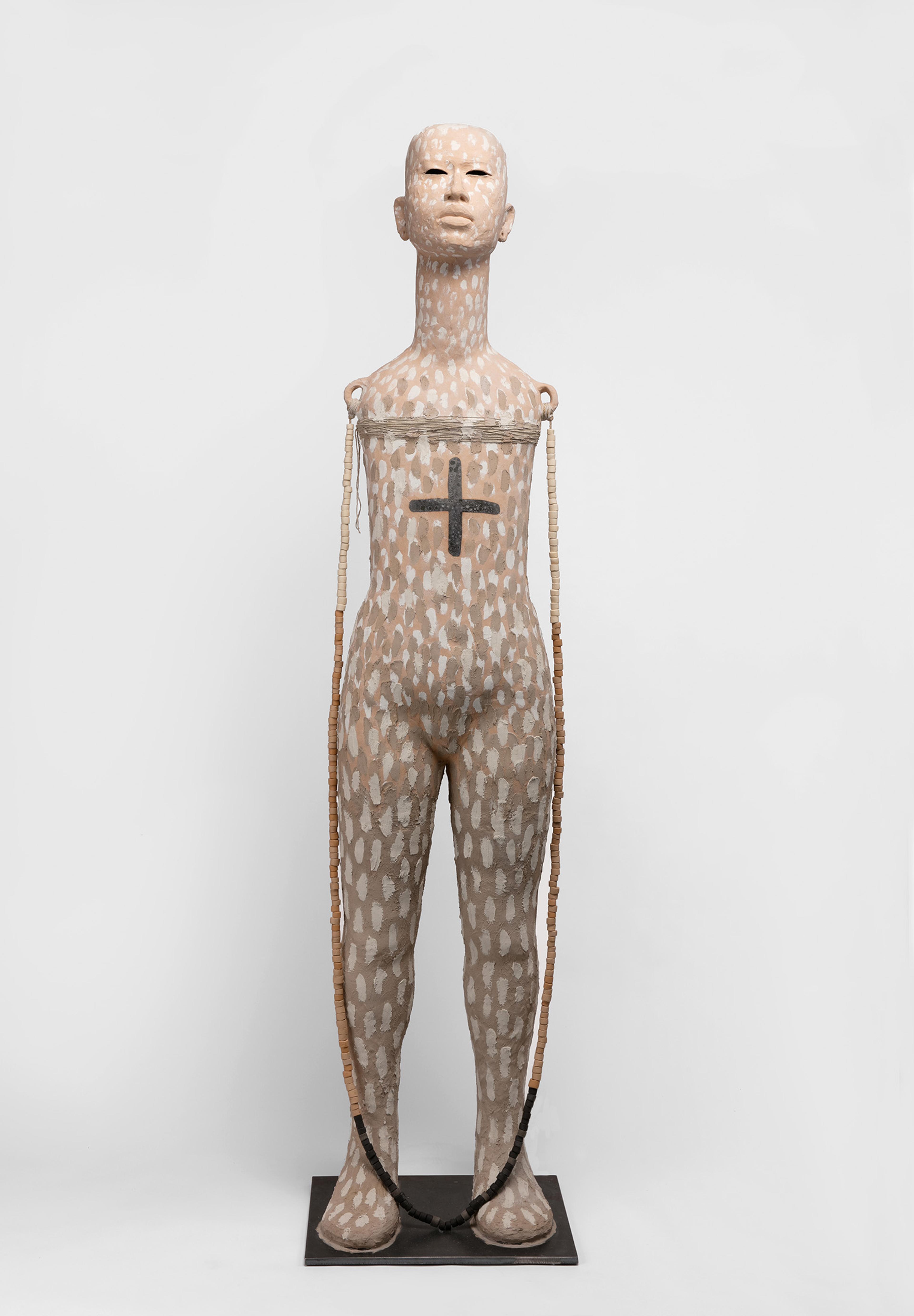 A tall, patterned sculpture depicts a human form with a prominent black cross on the chest. The figure's long neck and neutral expressio