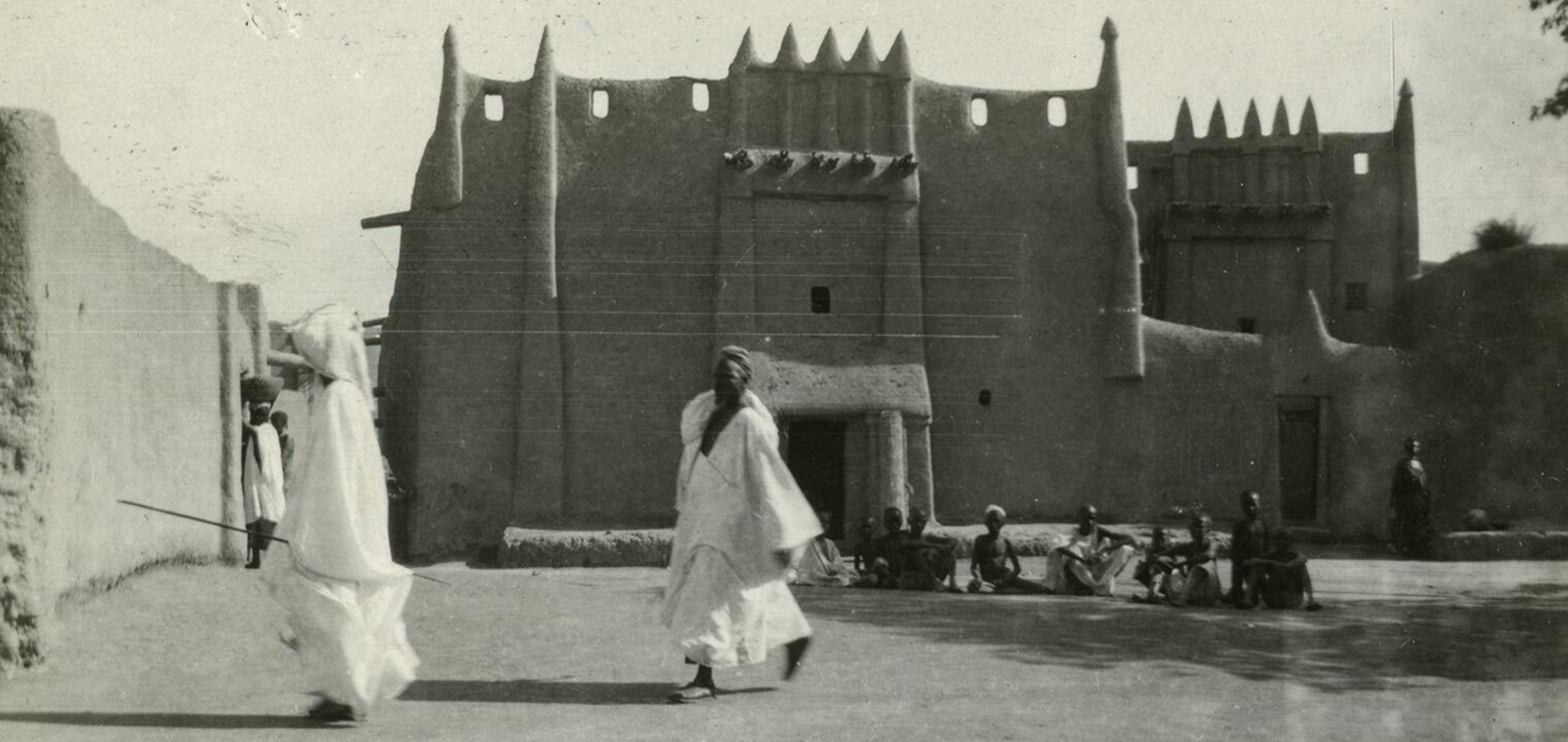 A black and white photo of two men in front of a mud building