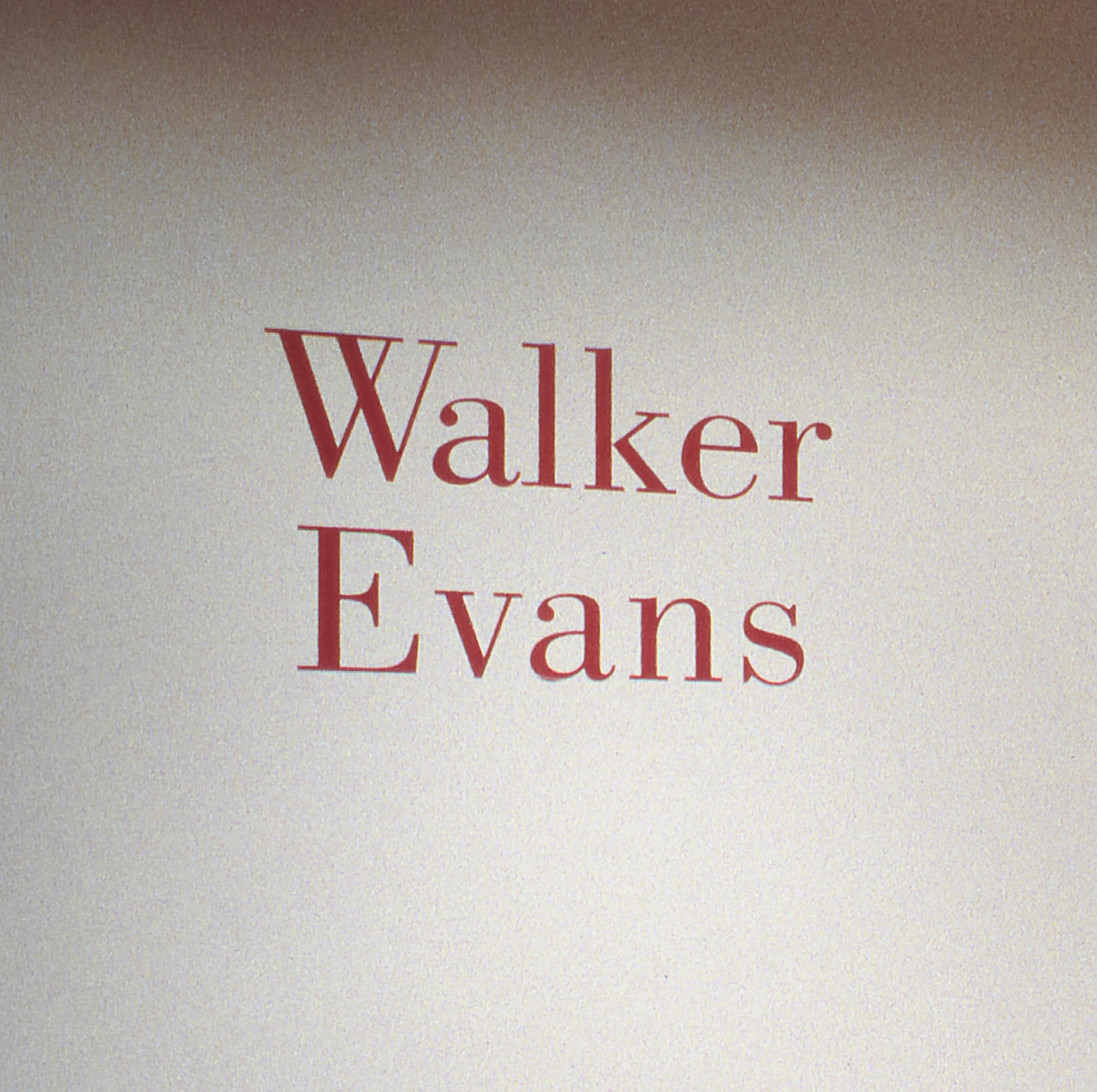 Red wall text reading "Walker Evans" against a white background, next to a gray wall panel with three frame photos.