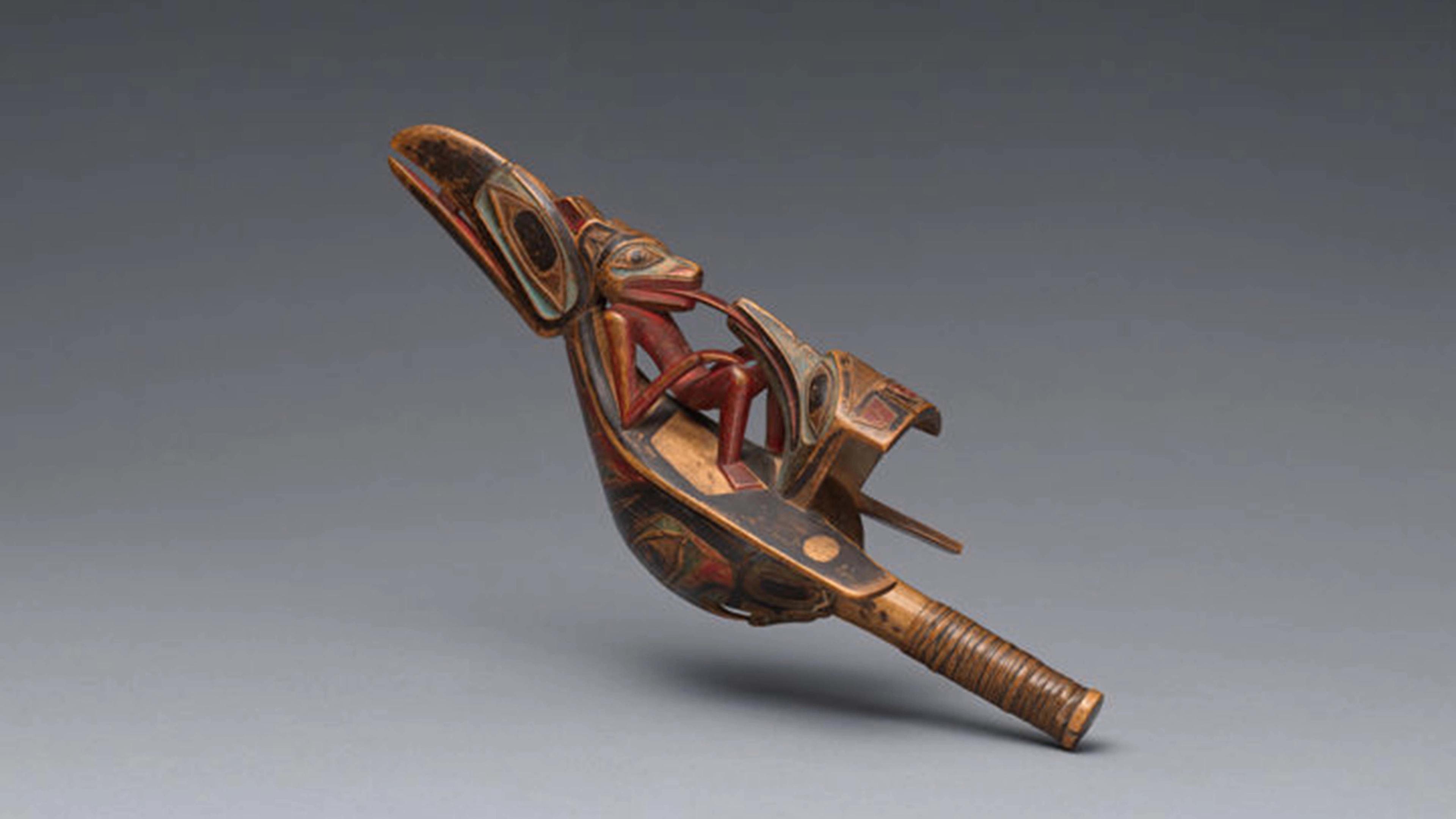 A carved wooden object featuring an intertwined human and animal form, painted in earthy red, black, and brown tones in a Northwest Coast Indigenous style