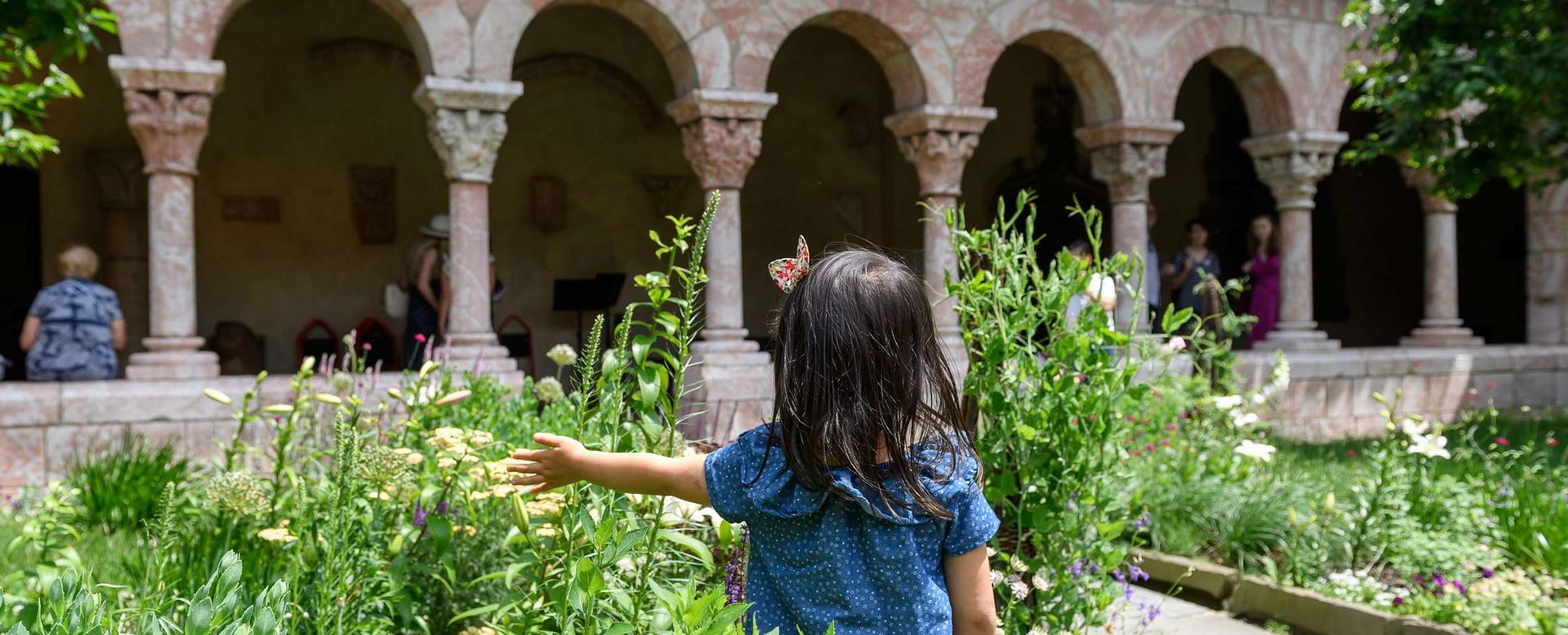 a young girl with her back turned to the camera stands in front of a garden bed in a medieval cloister.
