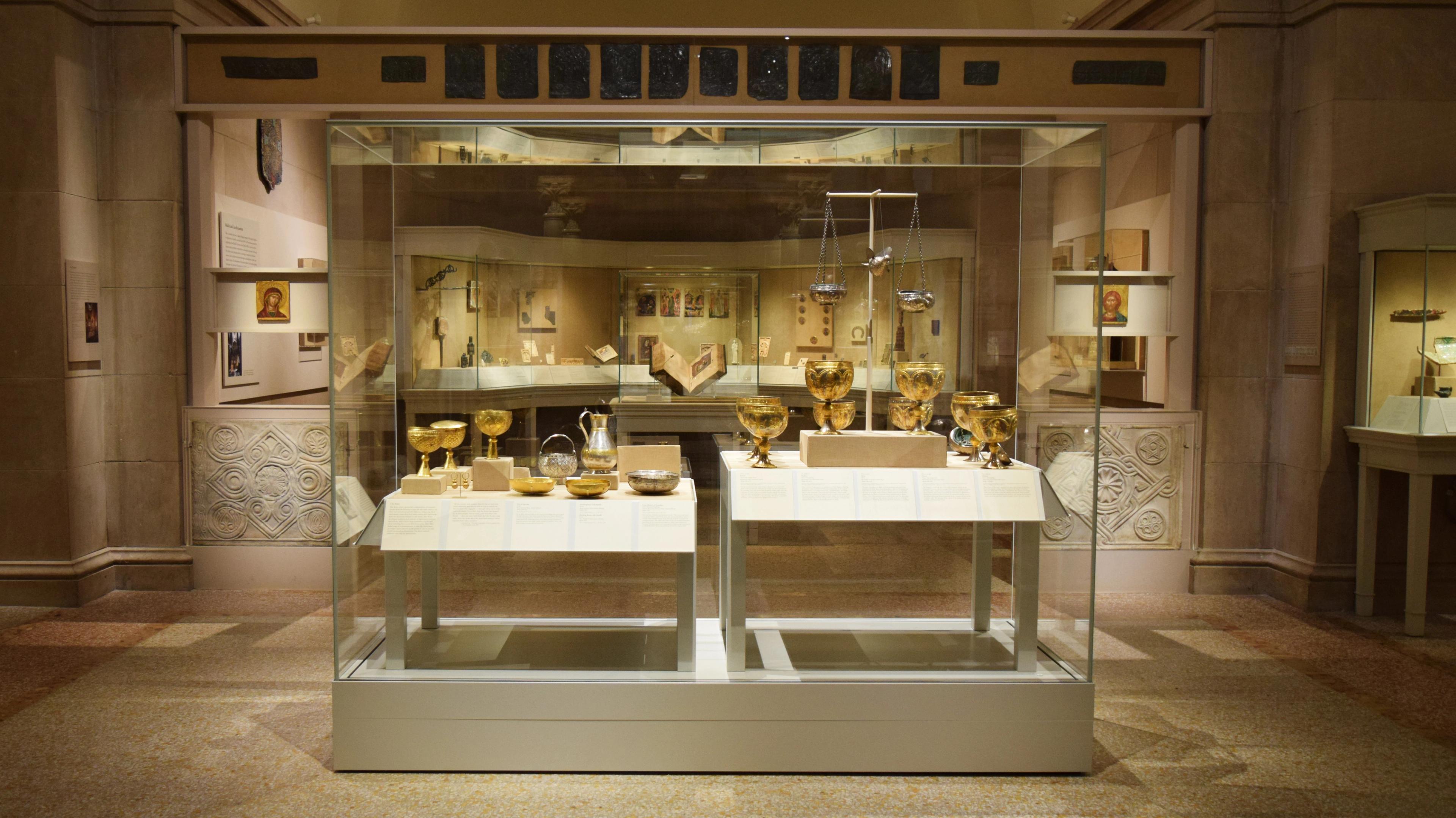 A display case in a large gallery holds several gold cups, bowls, and plates