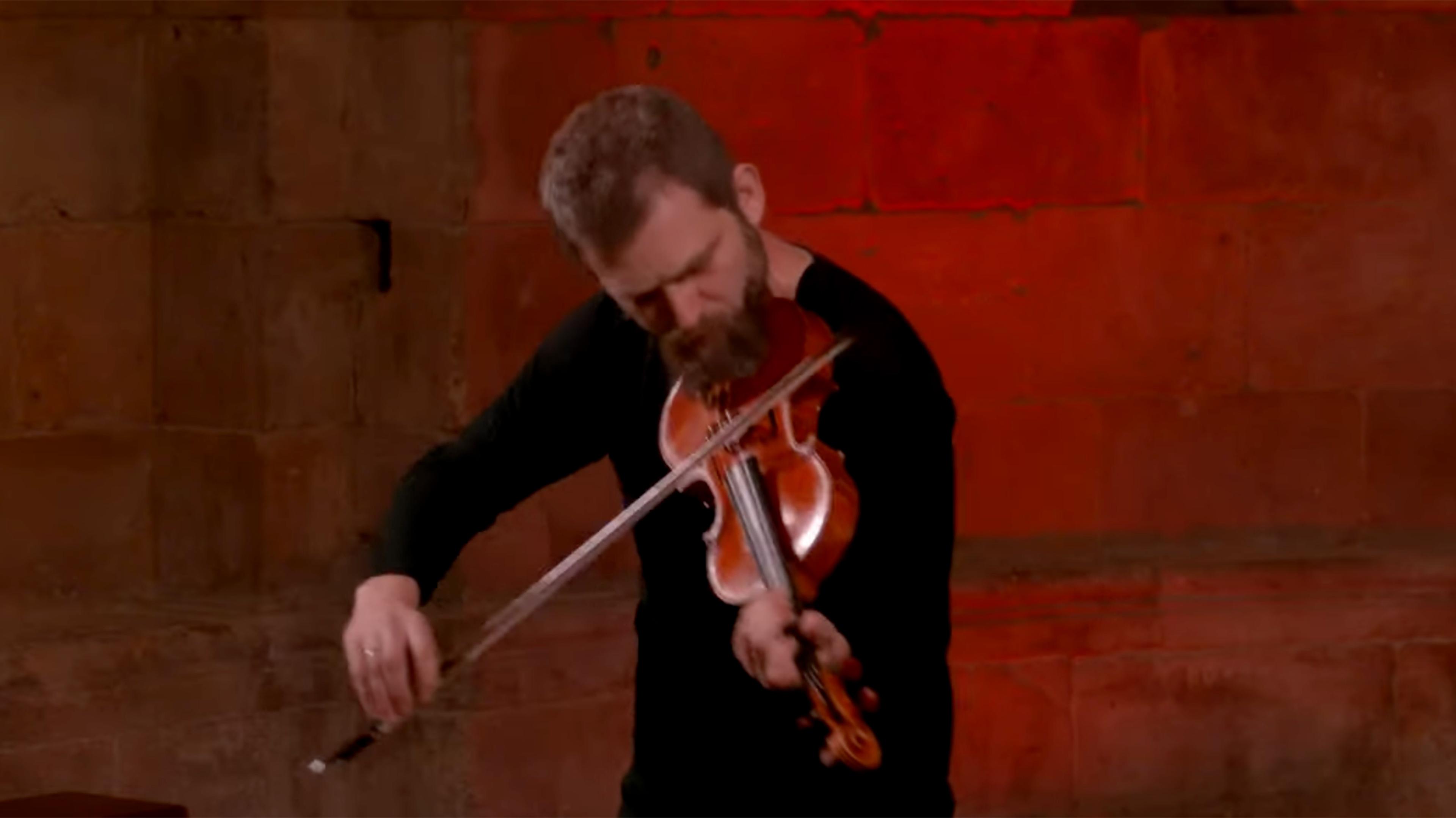 Johnny Gandelsman bent over a violin against a red-lit background.