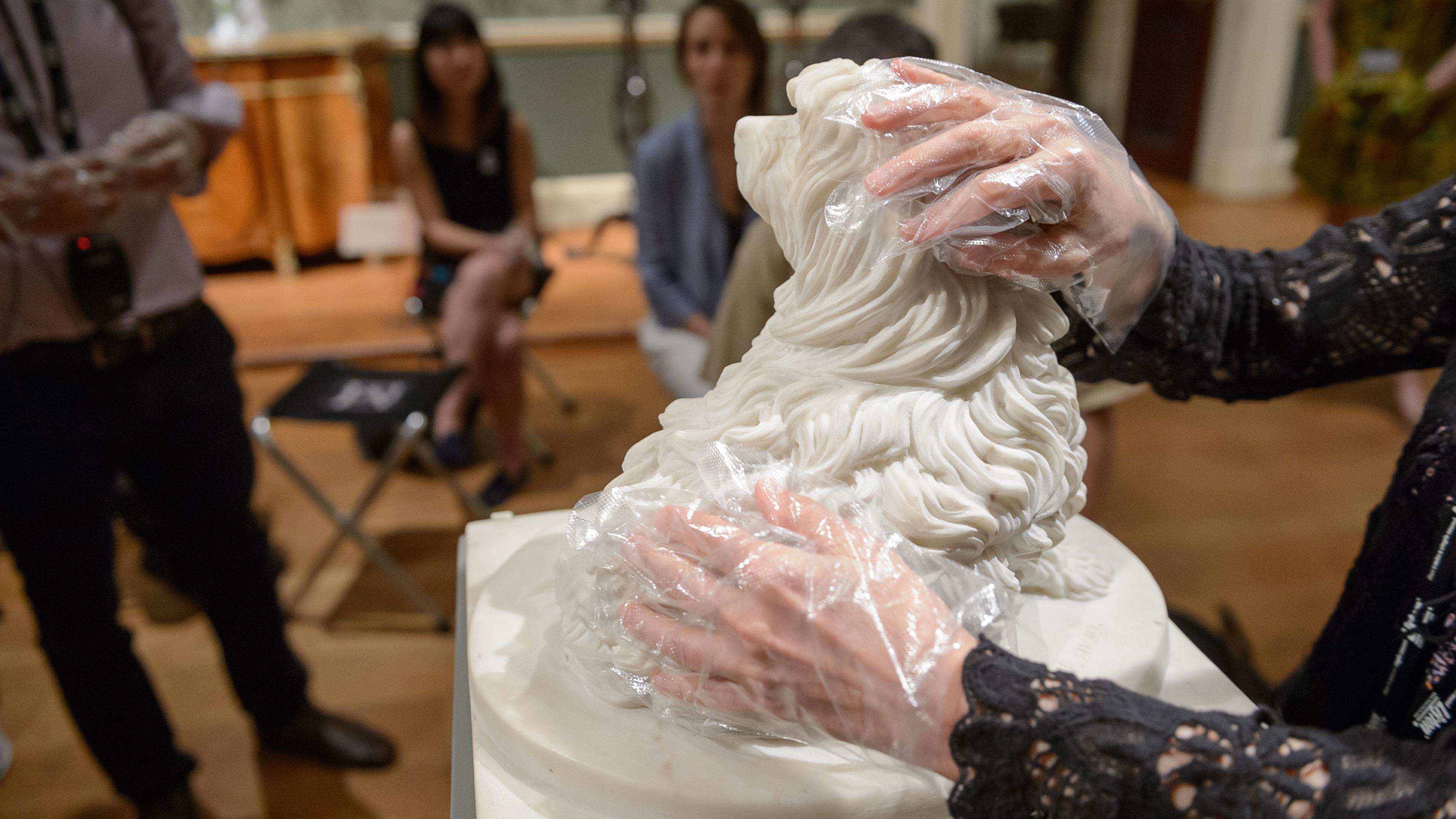 Accessibility. For visitors who are blind or partially sighted touch tours are available. A woman wearing plastic gloves touches a plaster sculpture. 