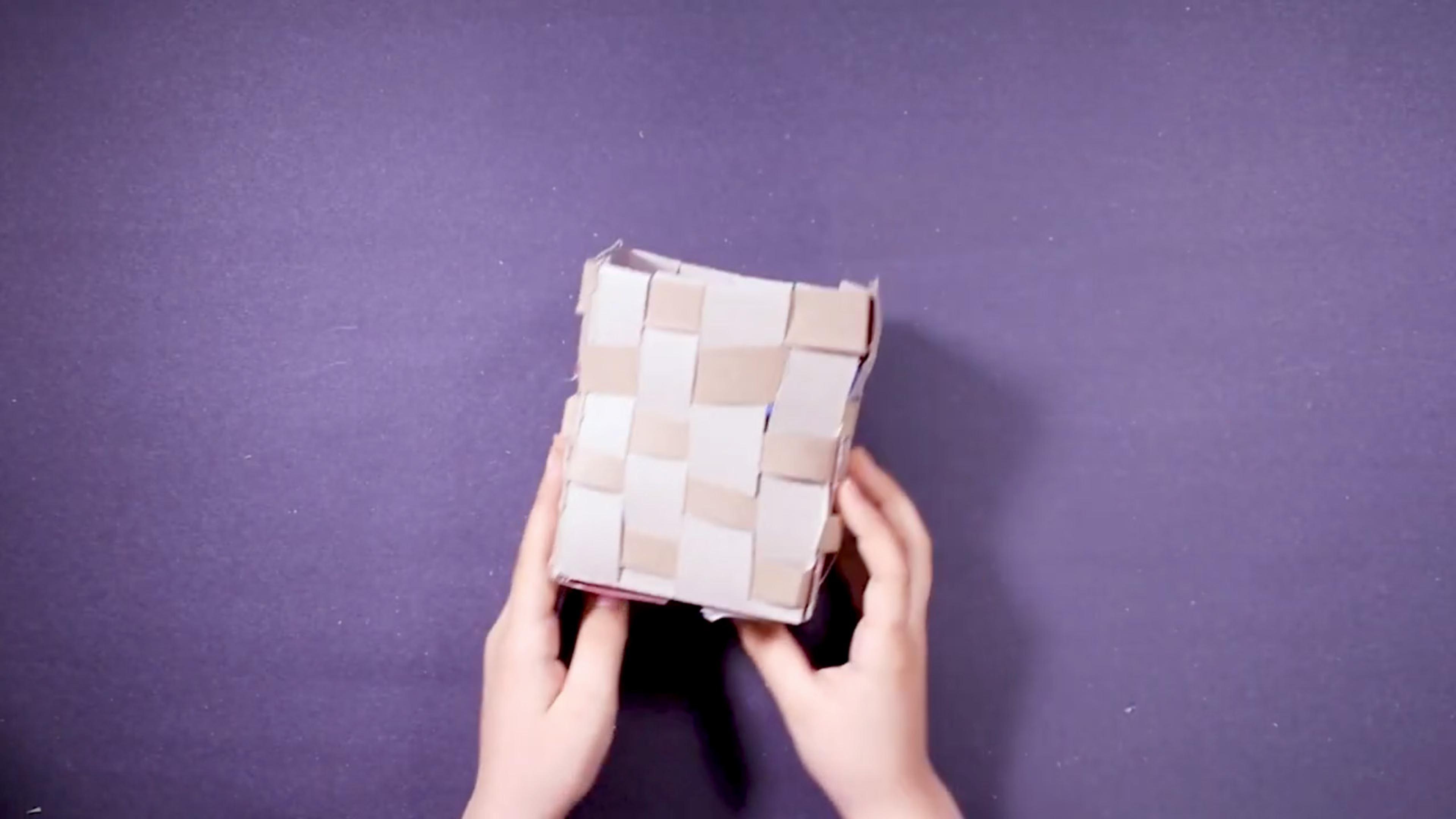 Hands holding a square basket made of woven paper against a purple background.