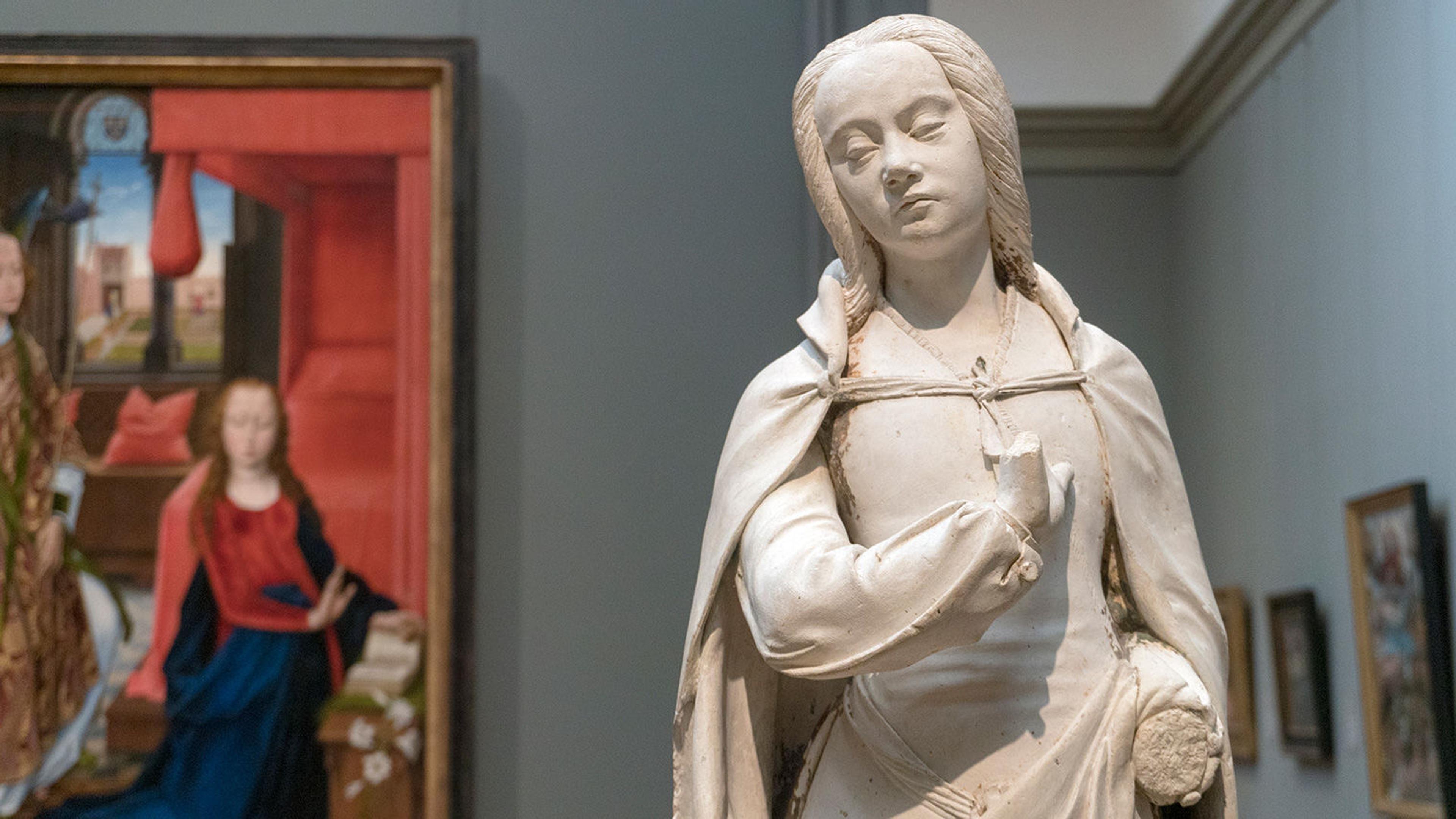 A closeup view of a statue of the Virgin Mary with a painting in the background