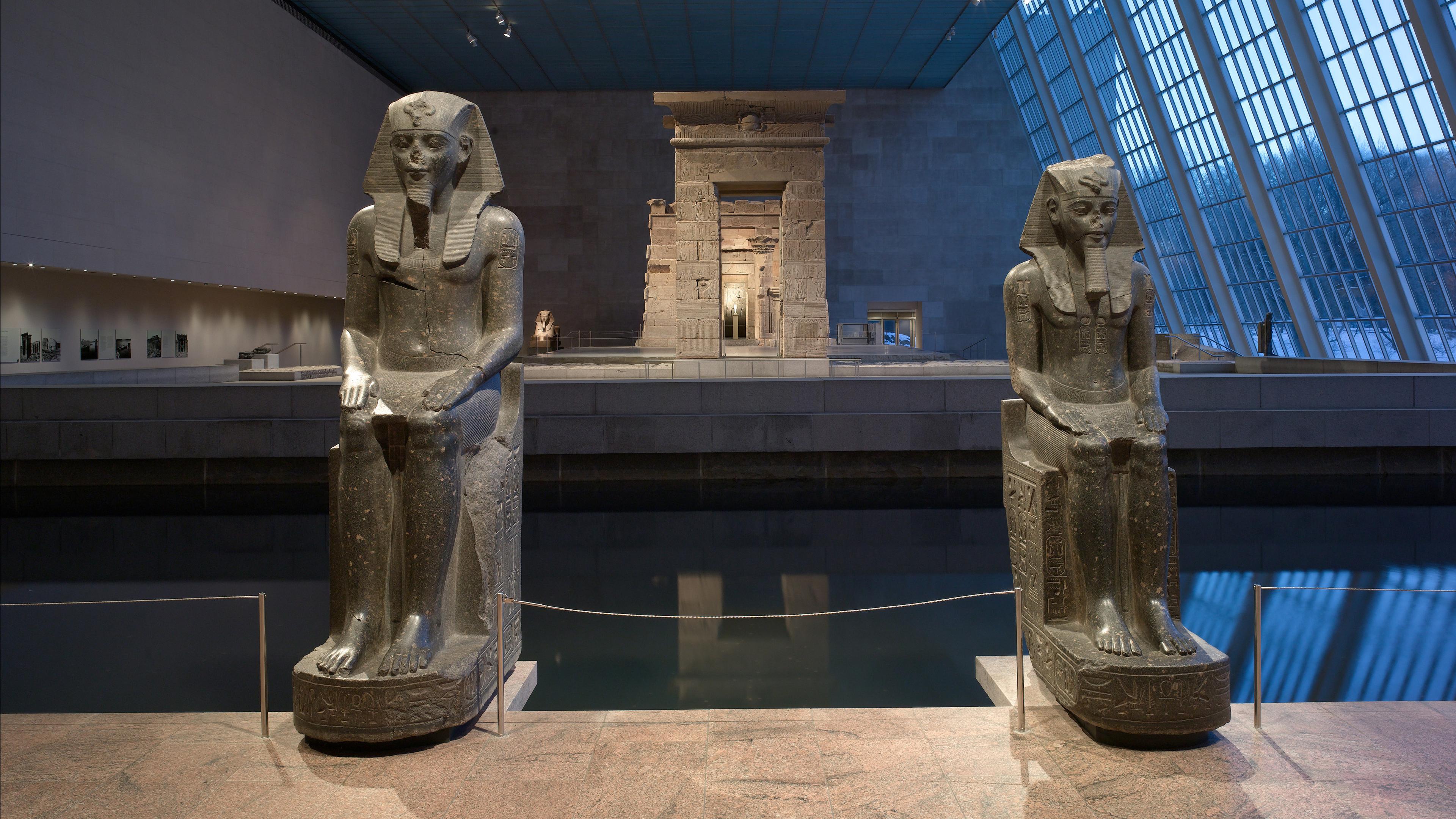 Two large seated statues of ancient Egyptian pharaohs flank a reflecting pool, with the Temple of Dendur in the background and light coming in from a tall wall of windows inside The Met.
