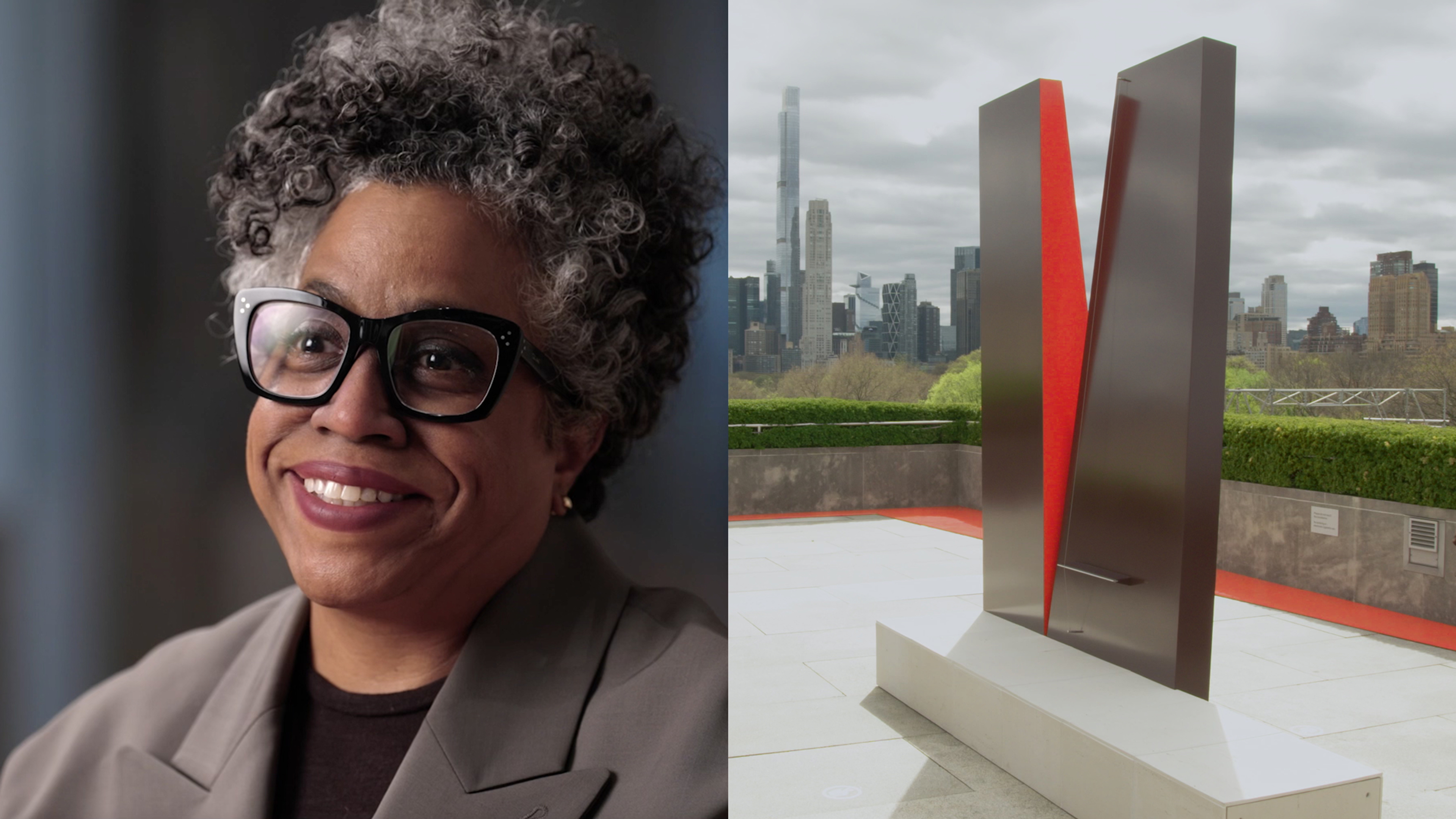 Portrait of a smiling person with gray curly hair and black glasses alongside a red and black abstract sculpture on a rooftop, with a city skyline in the background.