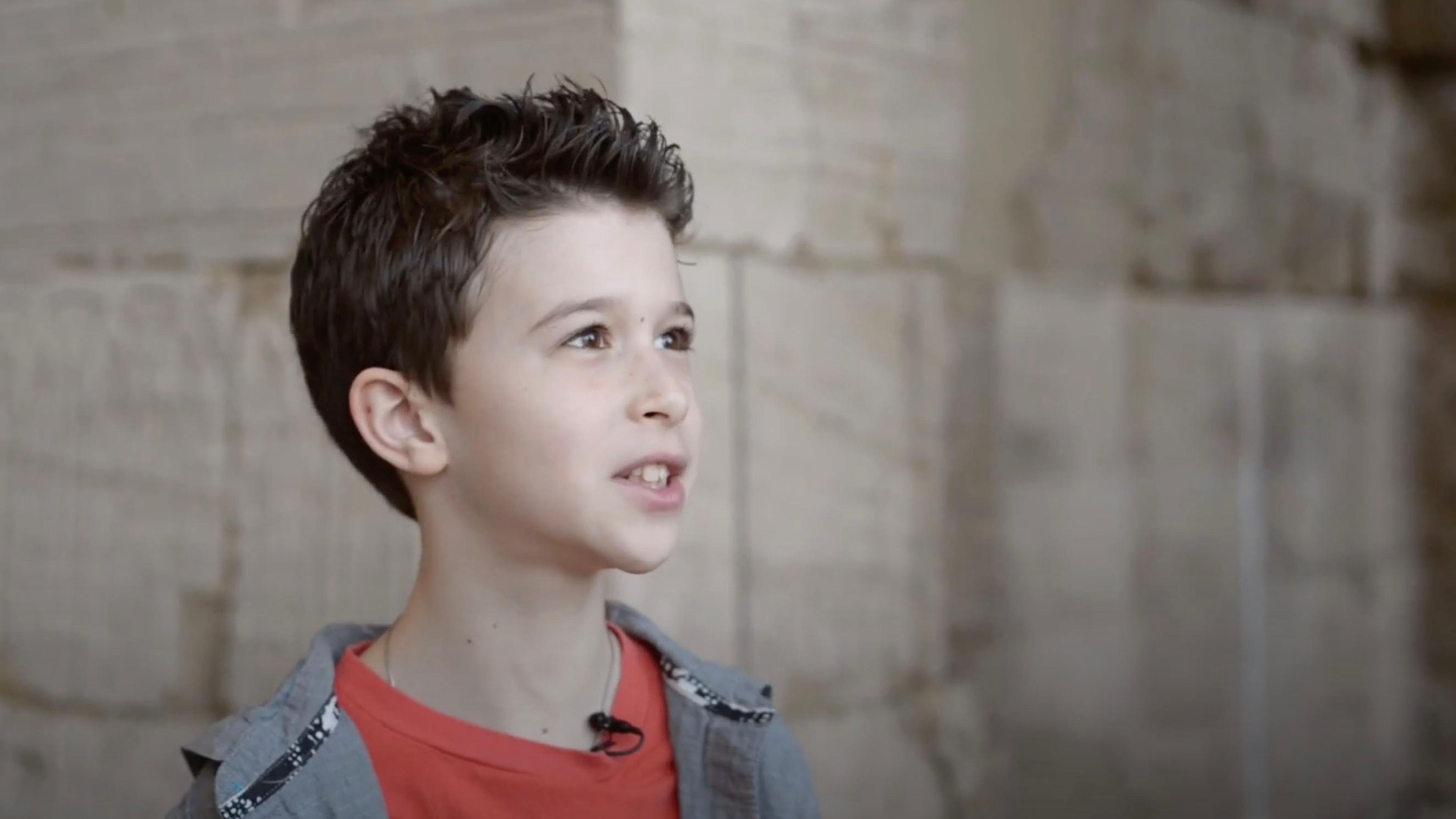 A young boy with dark, spiky hair and a red shirt is speaking while standing in front of a stone wall.