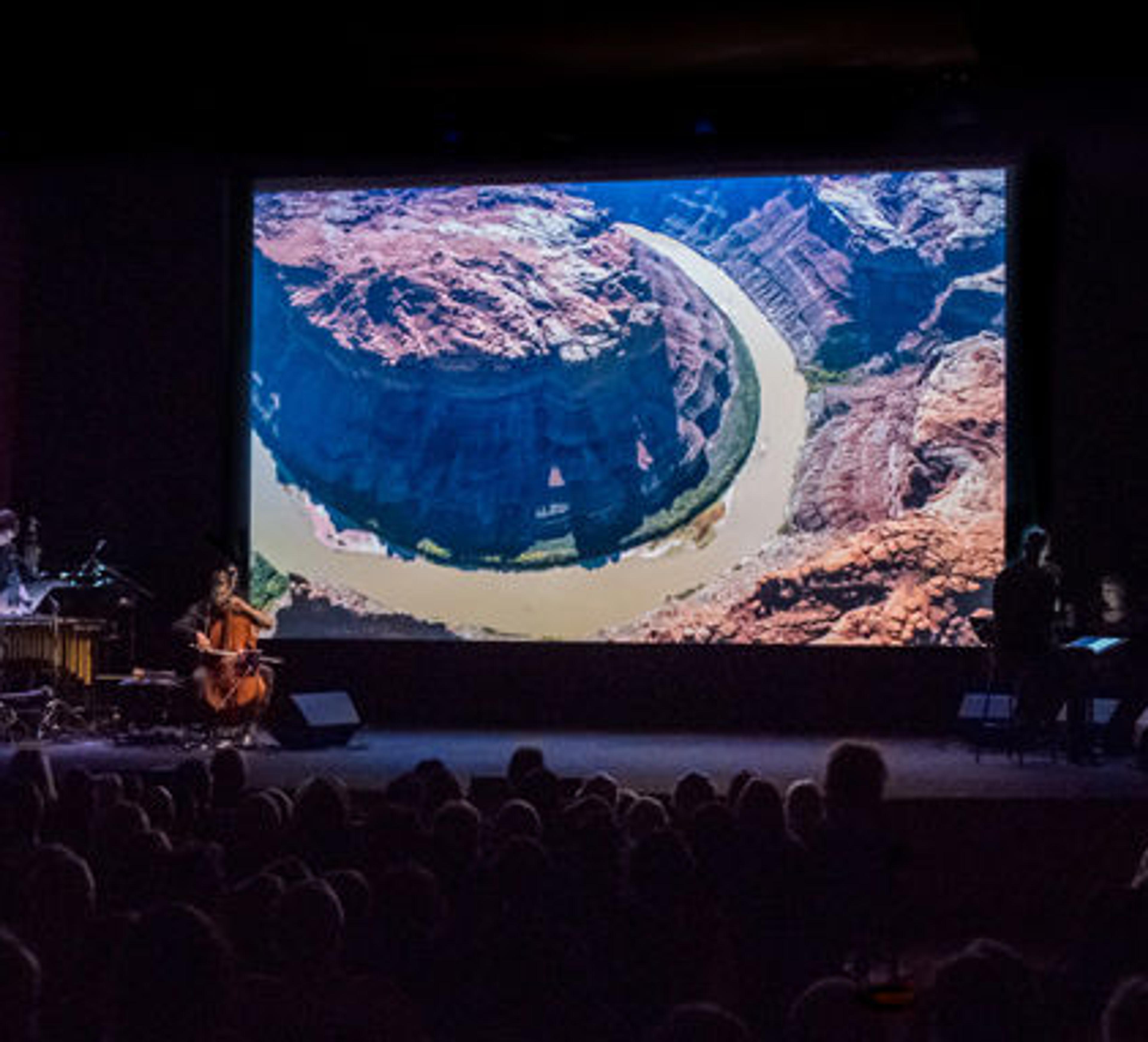 On a darkened stage several musicians and a chamber choir perform before a screen projection of a film depicting the Colorado River basin
