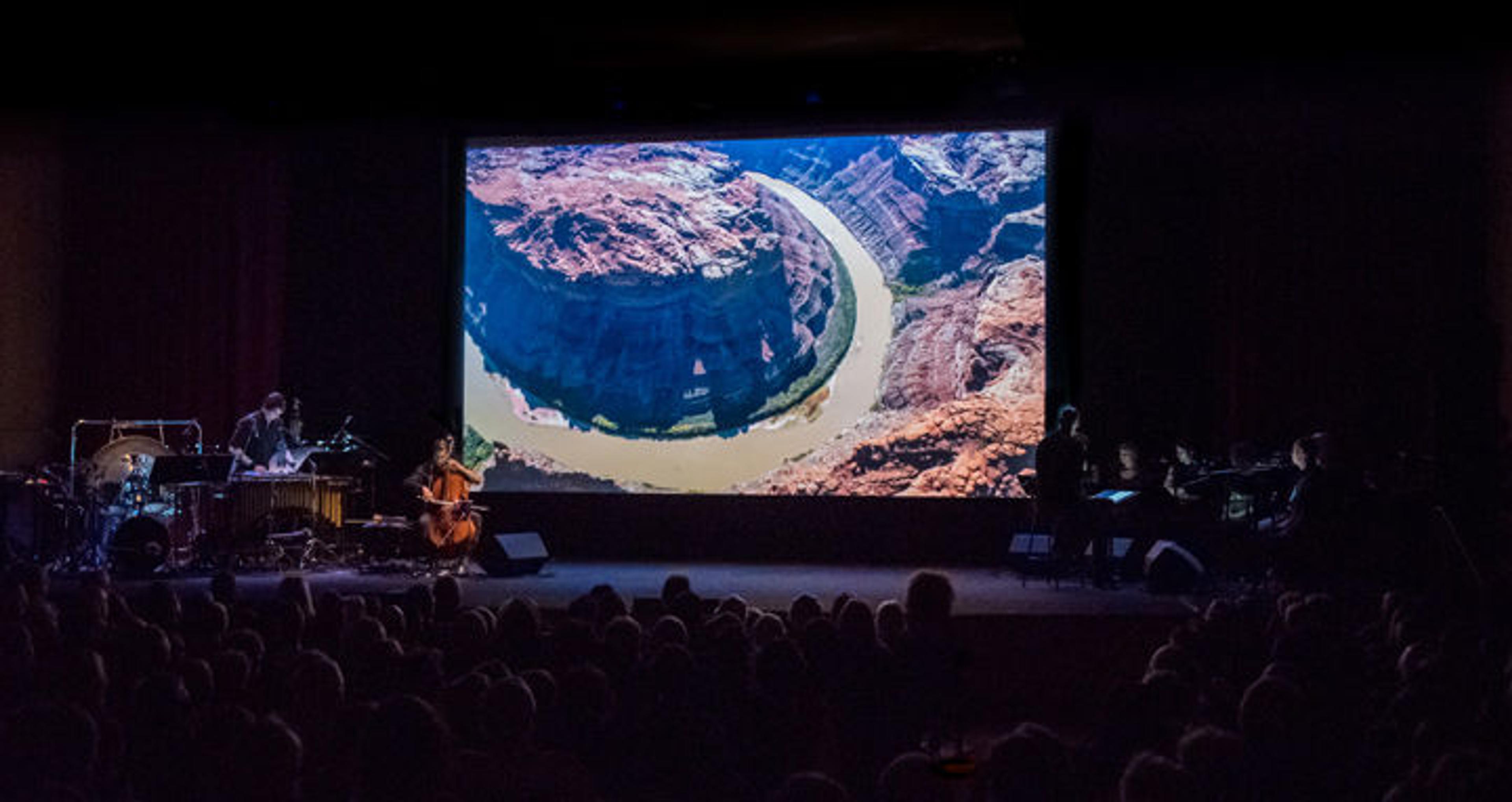 On a darkened stage several musicians and a chamber choir perform before a screen projection of a film depicting the Colorado River basin