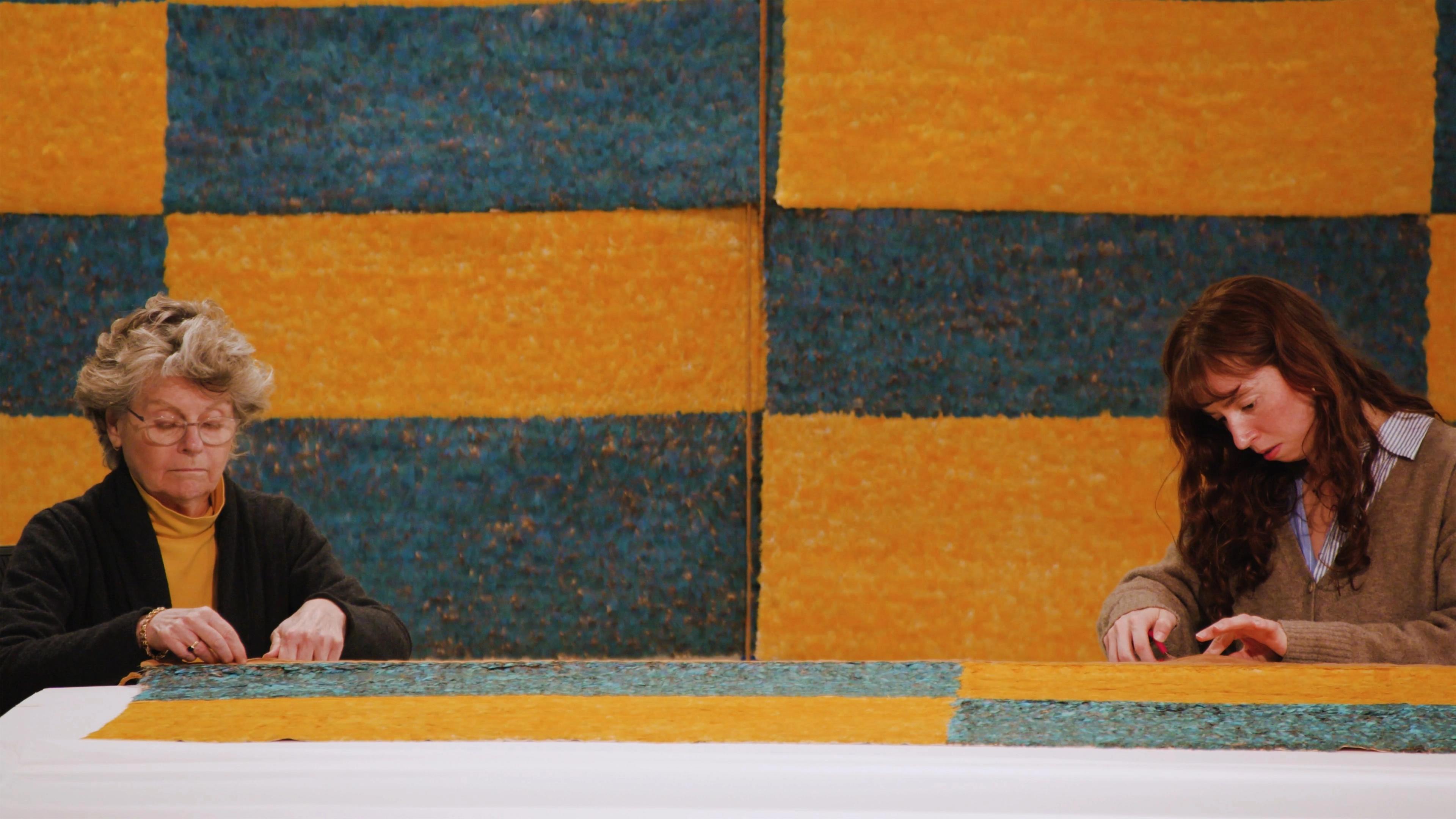 Two conservators working on preparing a Wari Feathered Panel for installation against a backdrop of checkered yellow and blue panels.