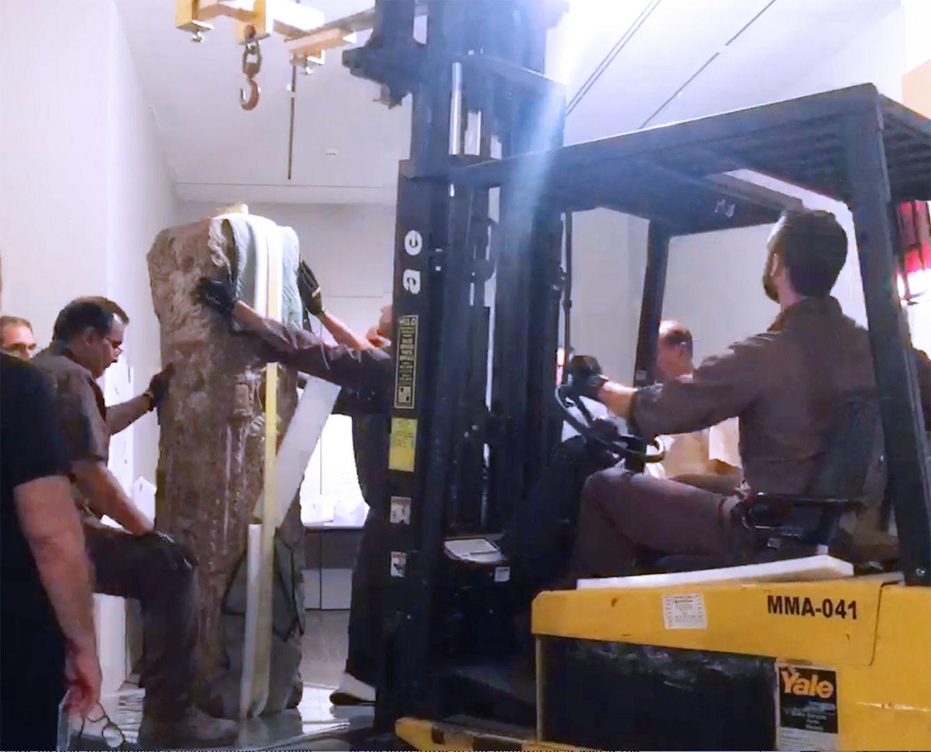 Men with yellow machinery hoisting a fifth-century stone stele in the galleries