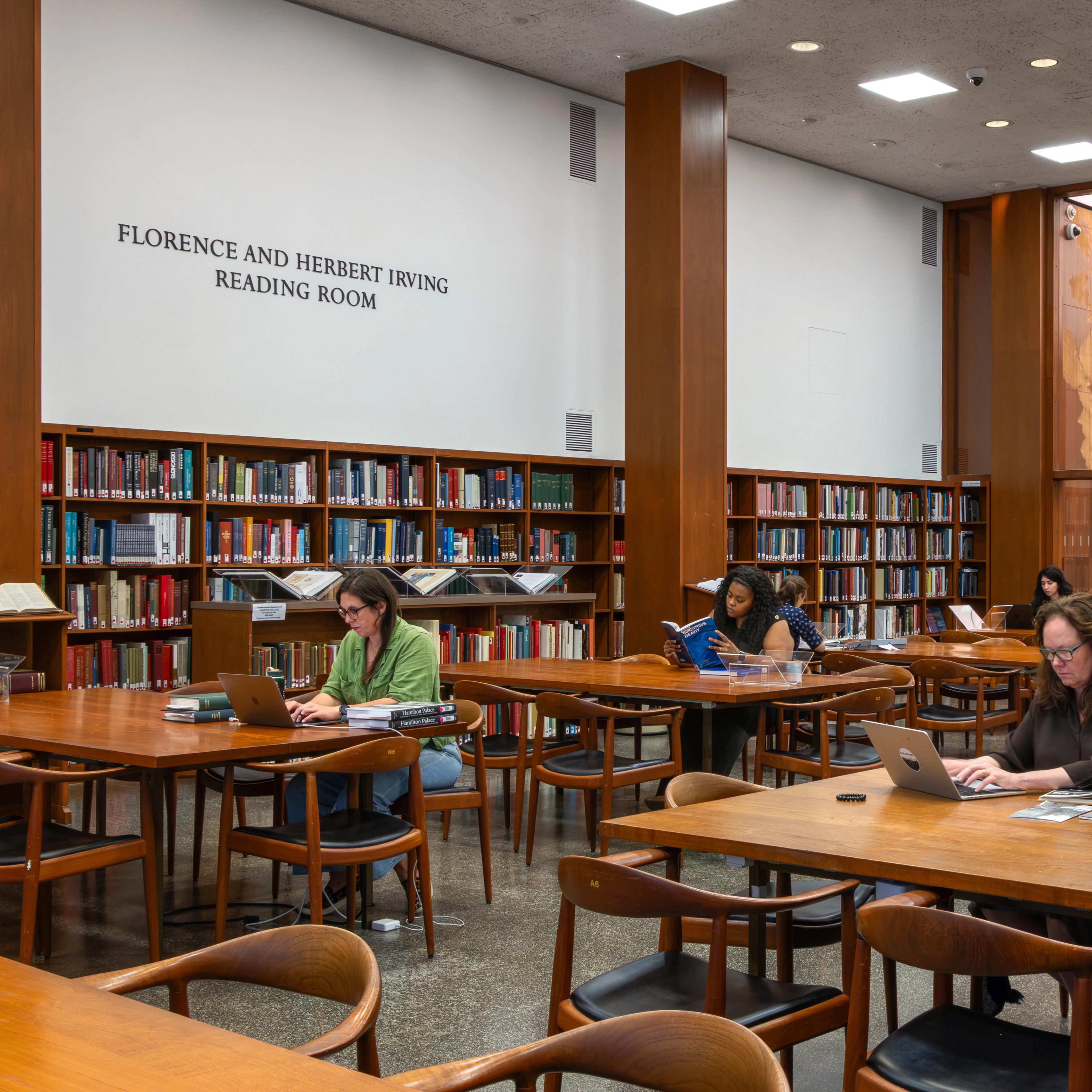 Reading room with bookshelves and study tables; visitors read and work on laptops beneath a sign reading “Florence and Herbert Irving Reading Room.”