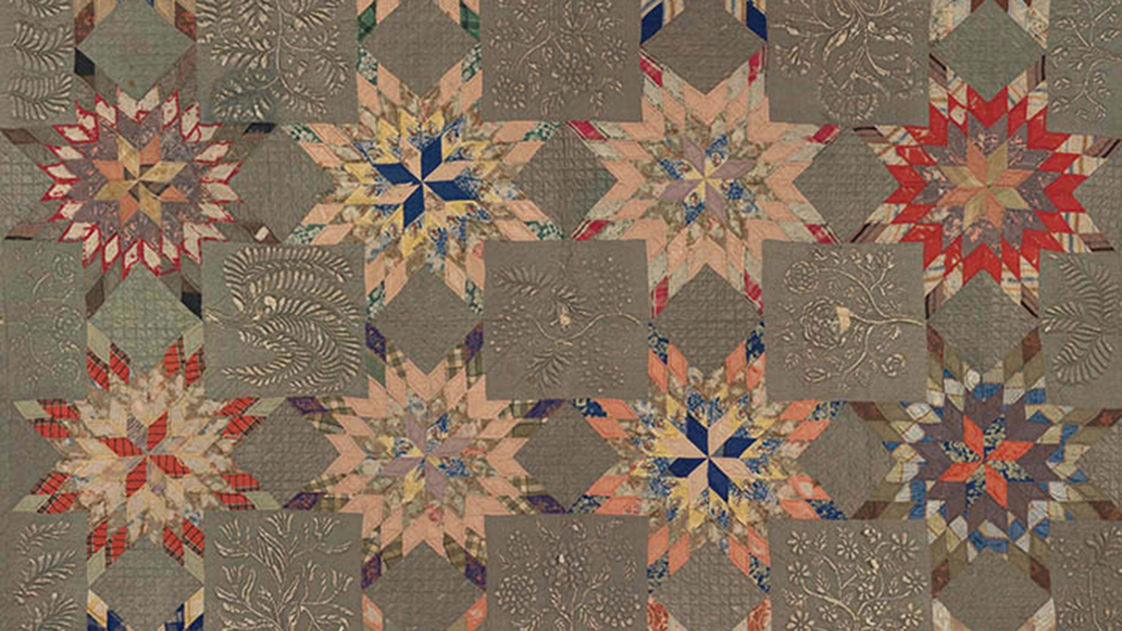 A quilt made of repeating star-shaped patchwork blocks in muted reds, blues, and browns, with embroidered floral and leaf motifs in the surrounding squares.