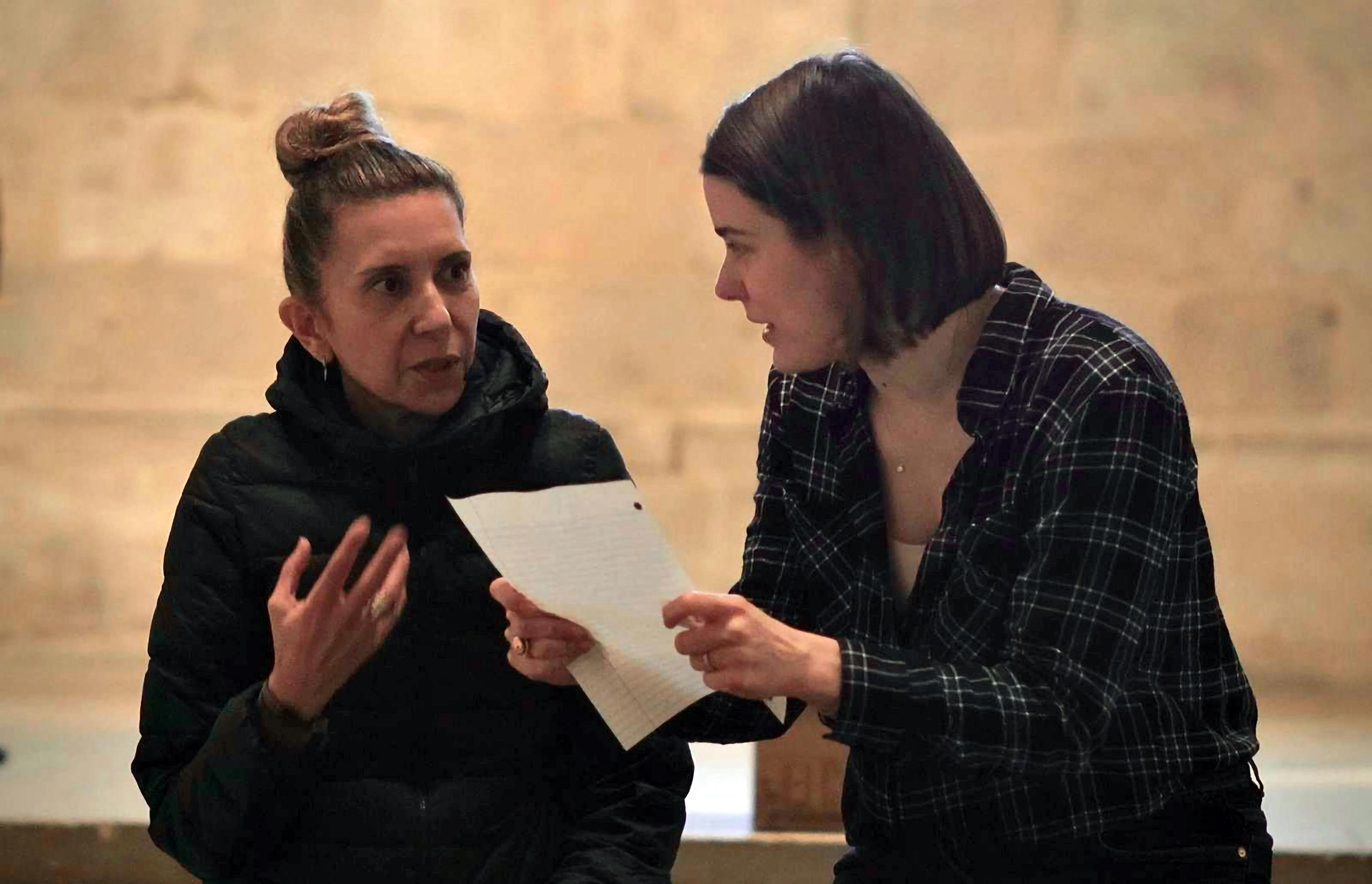 Two women angled towards each other, deep in discussion. The woman at right holds a sheet of paper.