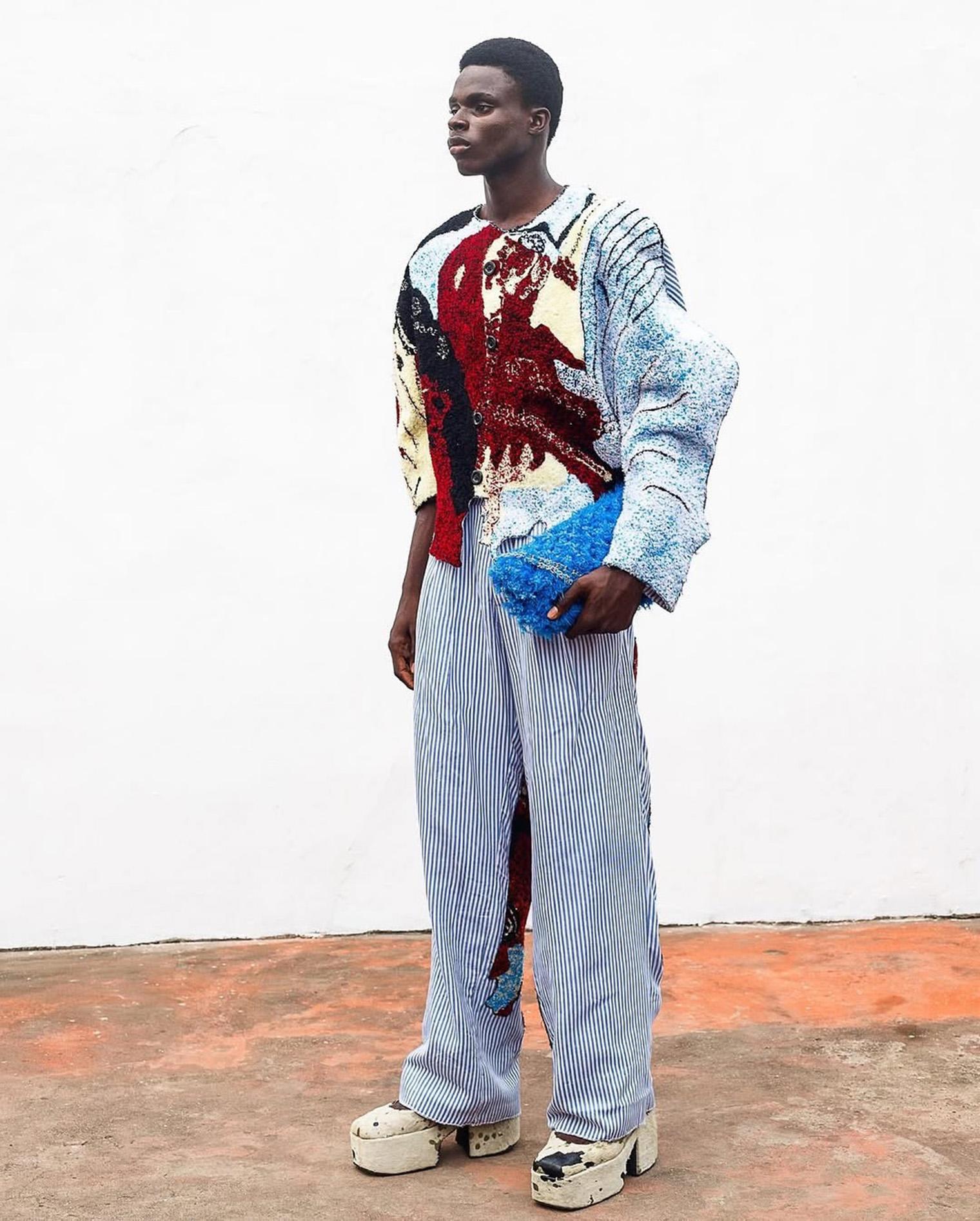 Young man in eclectic fashion: multicolored textured sweater, blue striped pants, platform shoes. Holds a fluffy blue clutch; neutral expression.