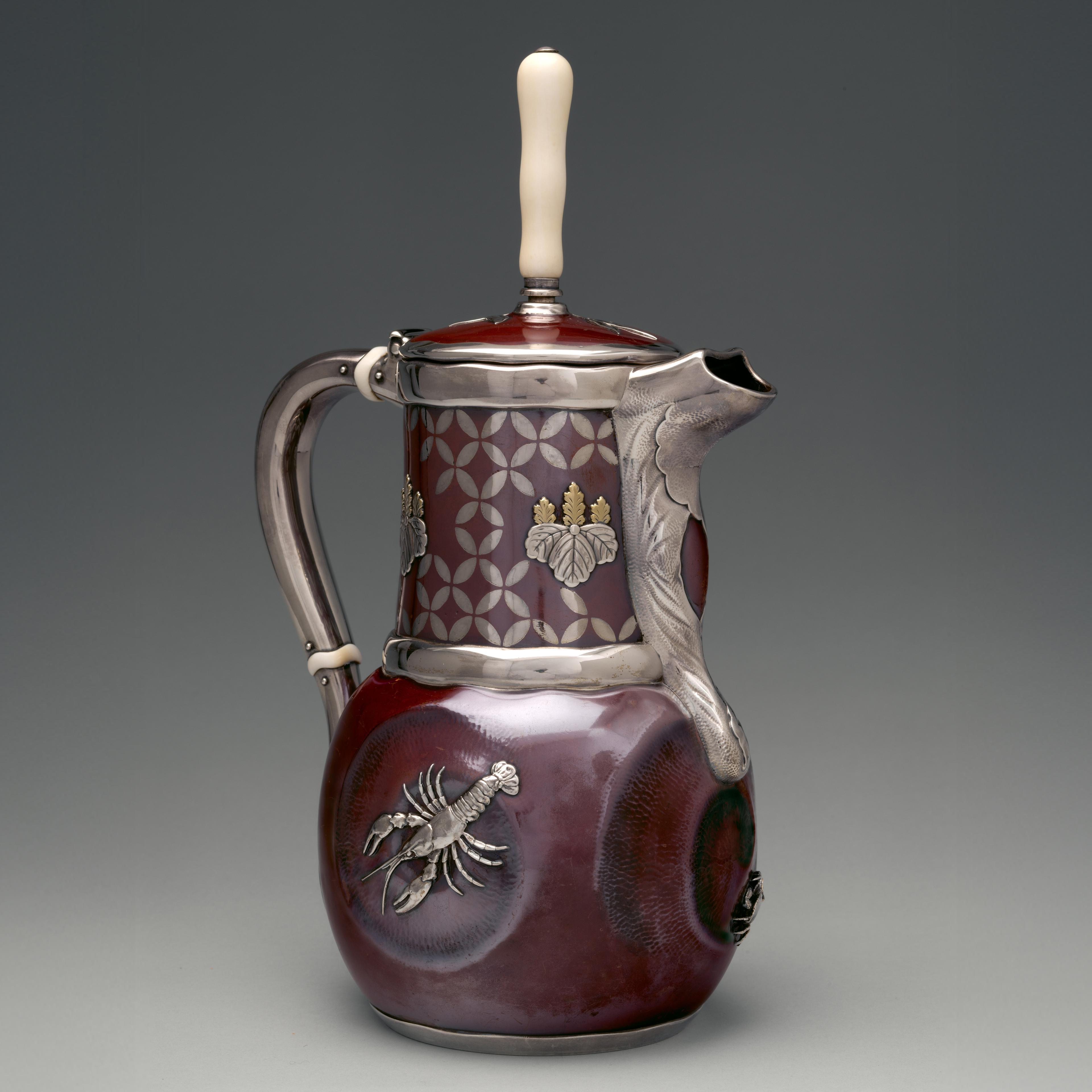 A three-quarter view of a patinated copper and silver chocolate pot in the shape of a pitcher with a handle, spot, and ivory handle. Silver banding emphasizes the handle, spout, and rim of the lid and neck. A lobster rendered in high-relief silver adorns the body of the pot.