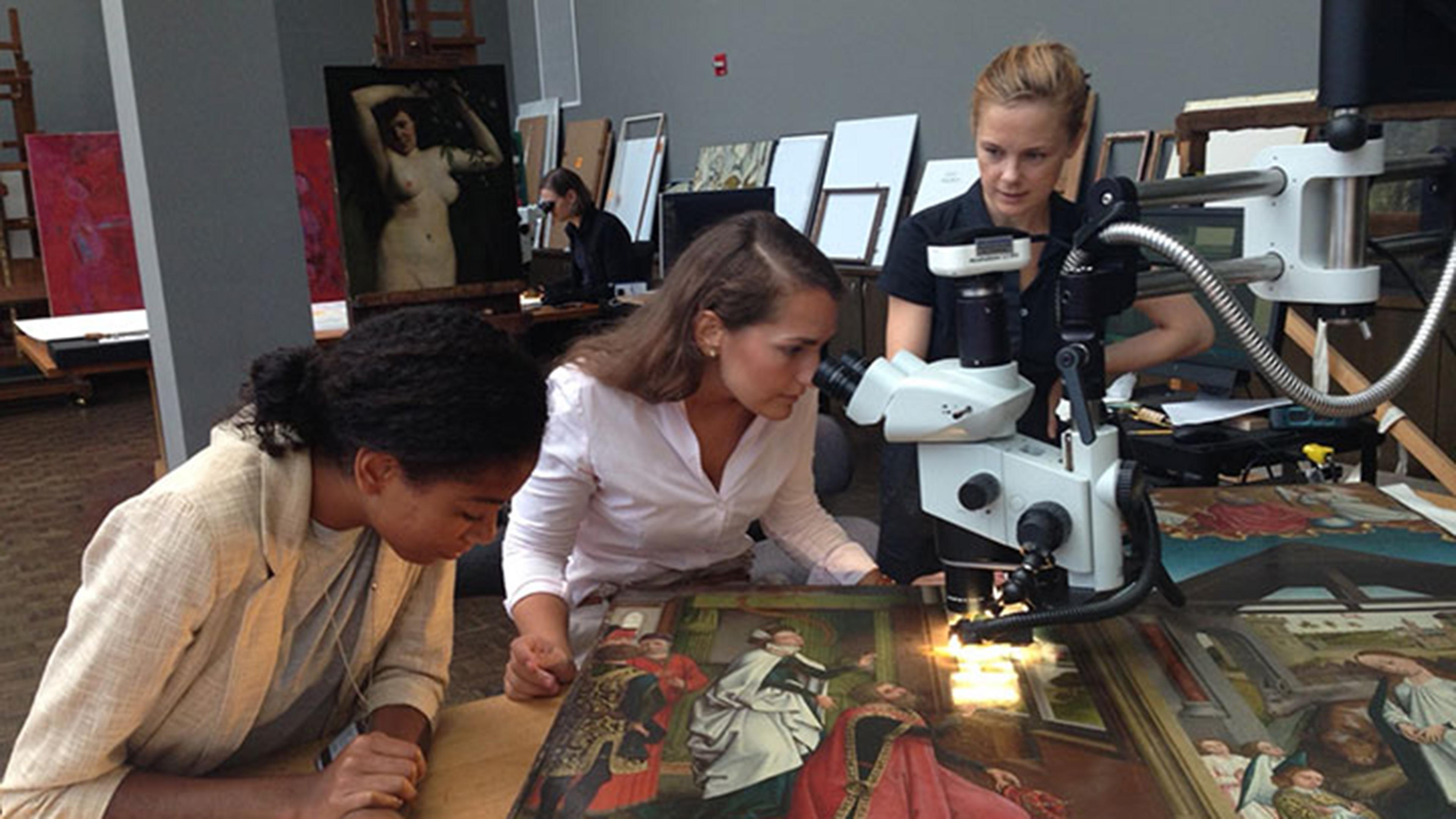 Conservator Sophie Scully and two curatorial fellows gather around a table in the Paintings Conservation studio. One of the fellows looks at a Flemish painting through a microscope while the others observe.