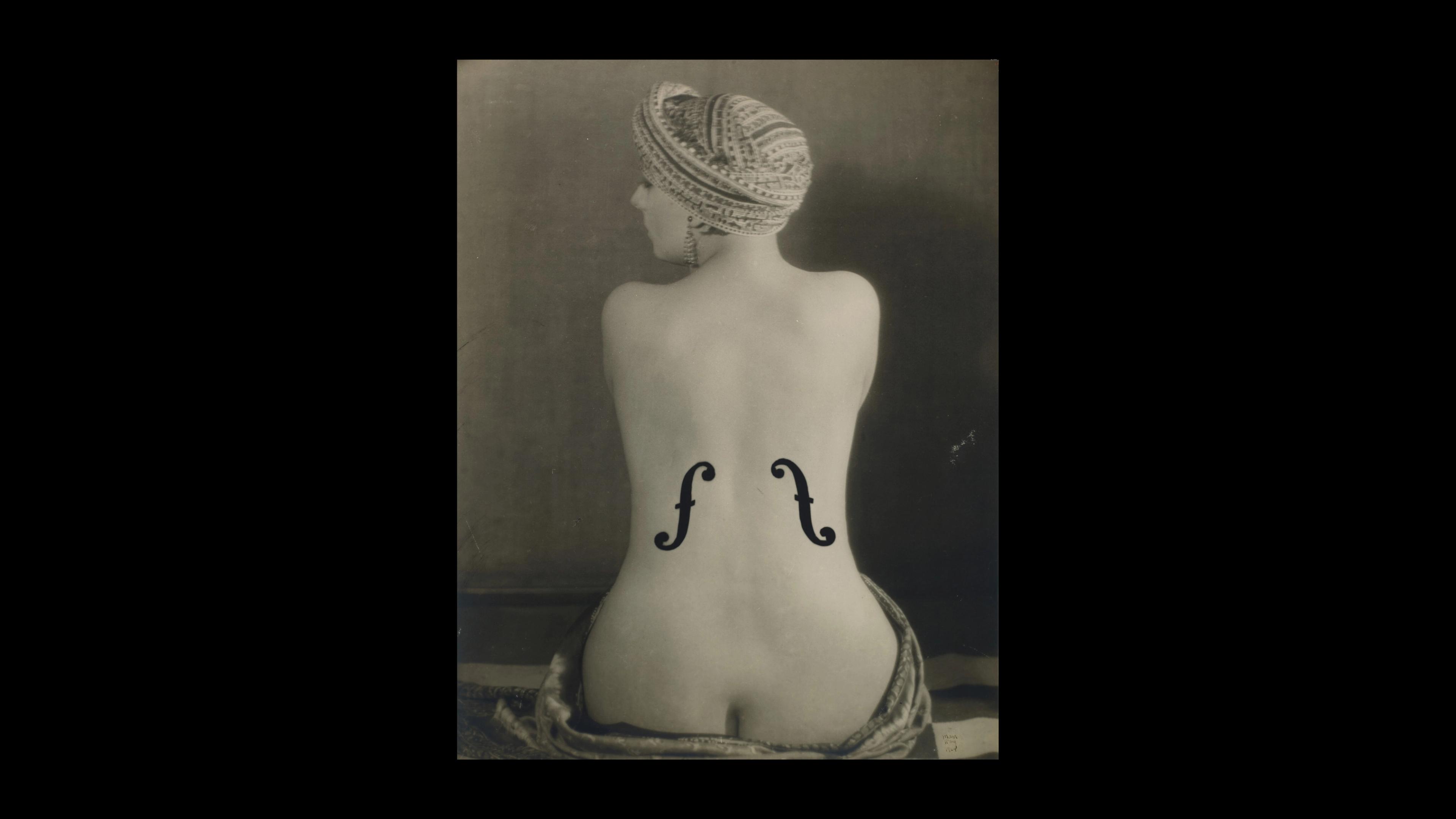 Black and white photograph showing the back of a seated nude person with a wrap around their head and waist with two stylized musical notes on the middle of the person's back.