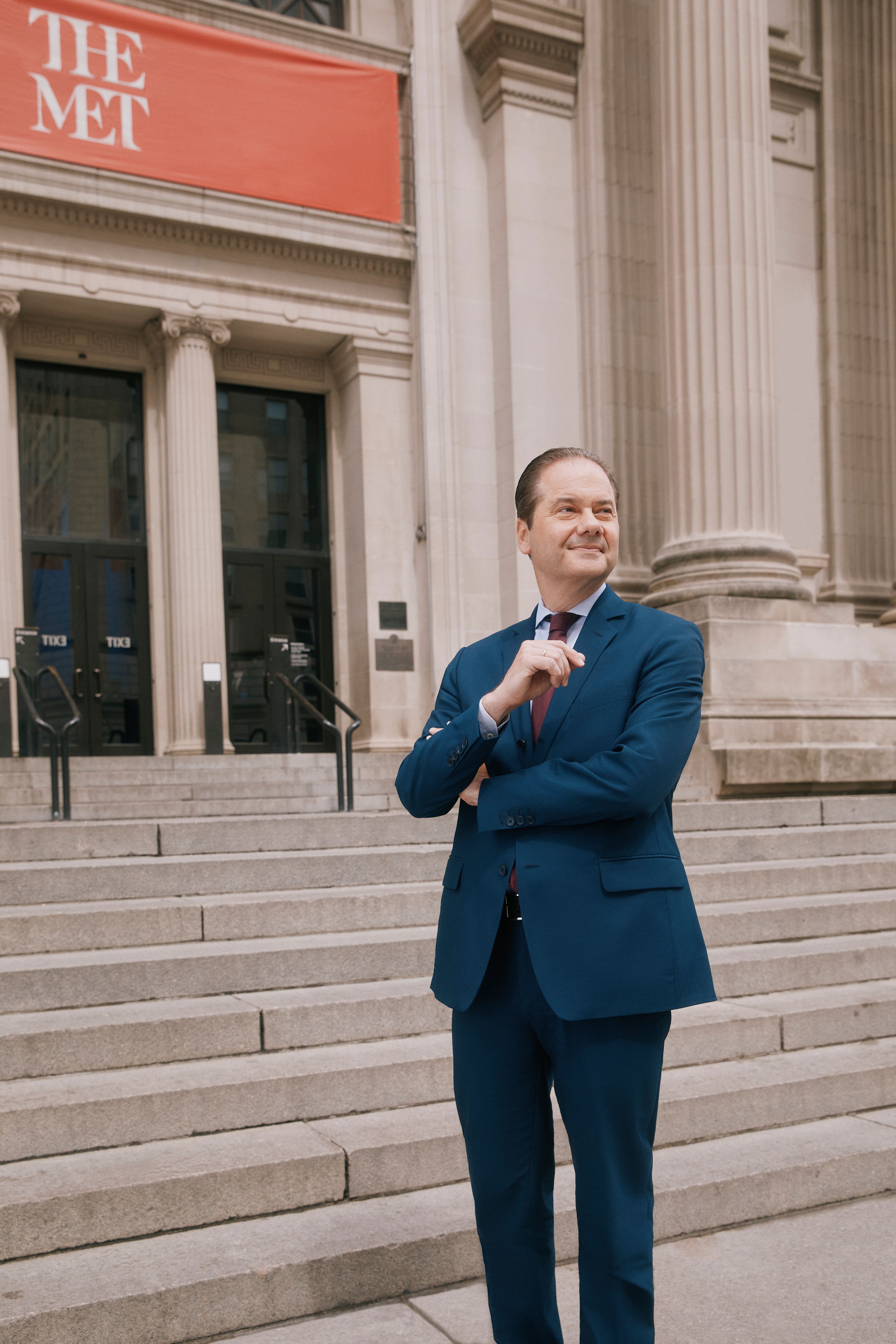 A color photograph of Max Hollein standing in a blue suit on the steps outside The Met Fifth Avenue entrance.