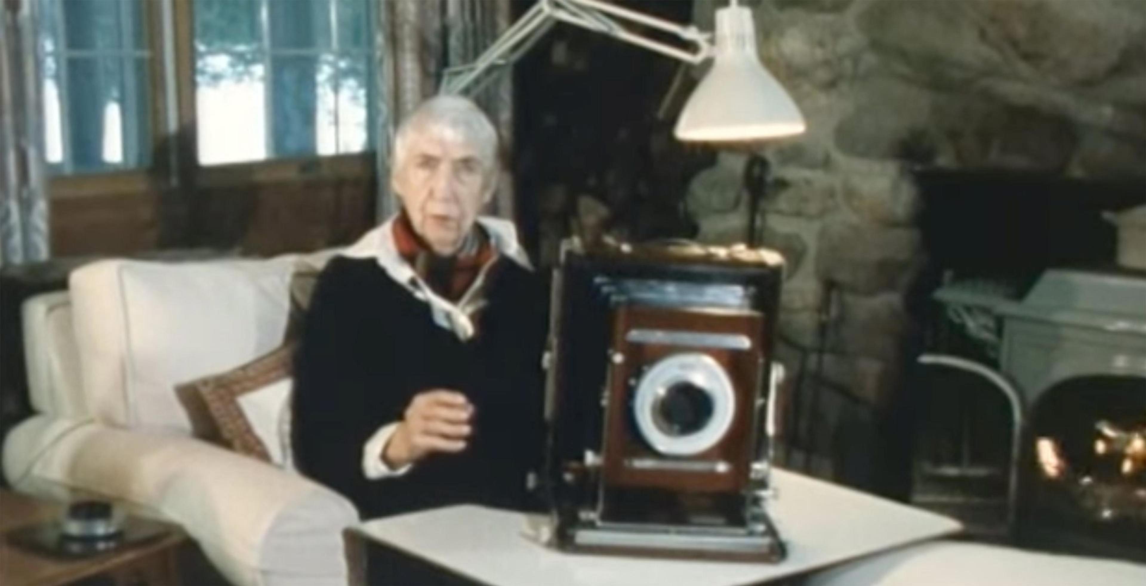Still from "The Living Room" of Berenice Abbott sitting in an armchair in front of a fire with a camera on the table in front of her