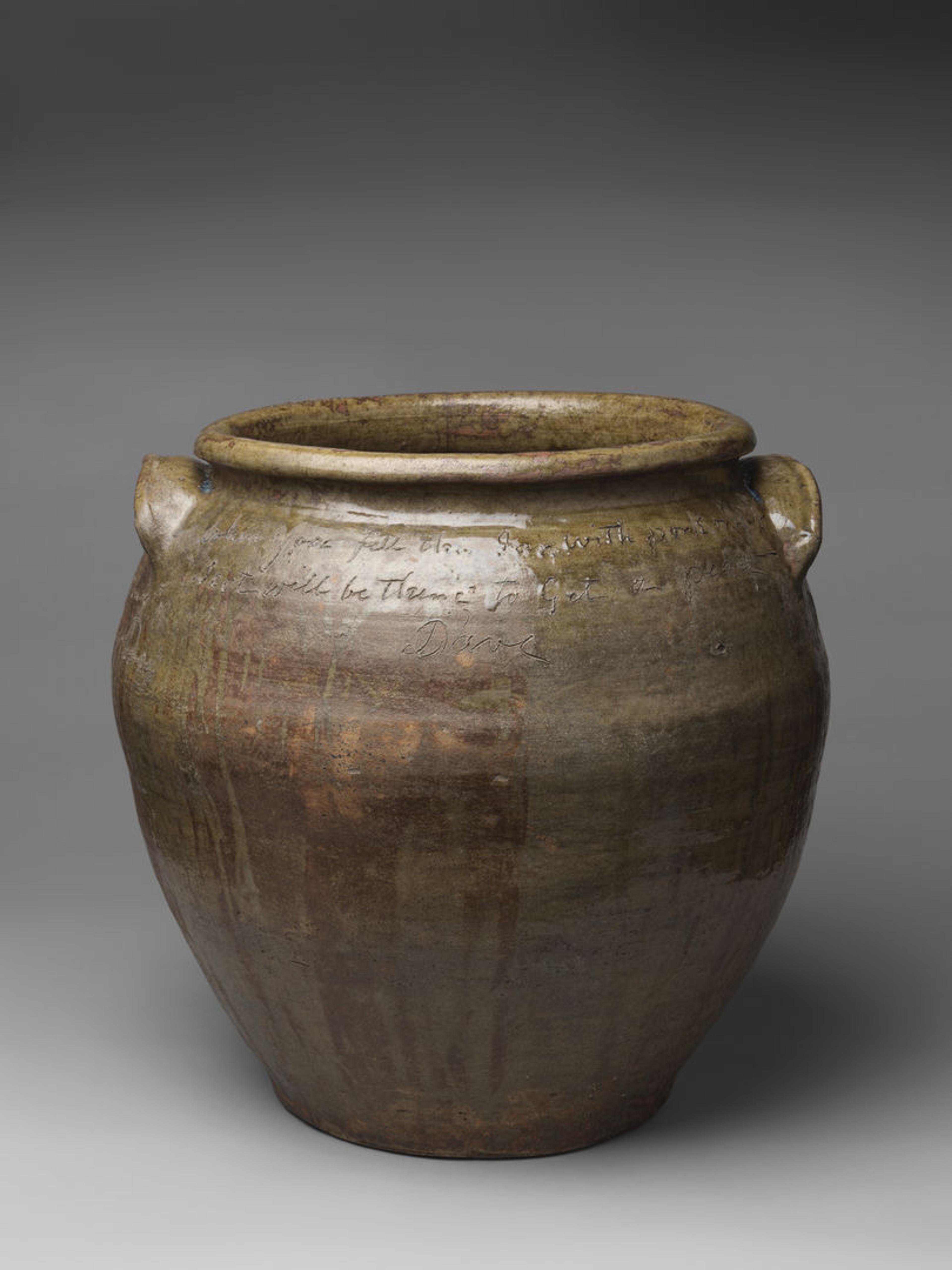 Photograph of brownish yellow stone storage jar with text inscription on its surface, against a grey backdrop.