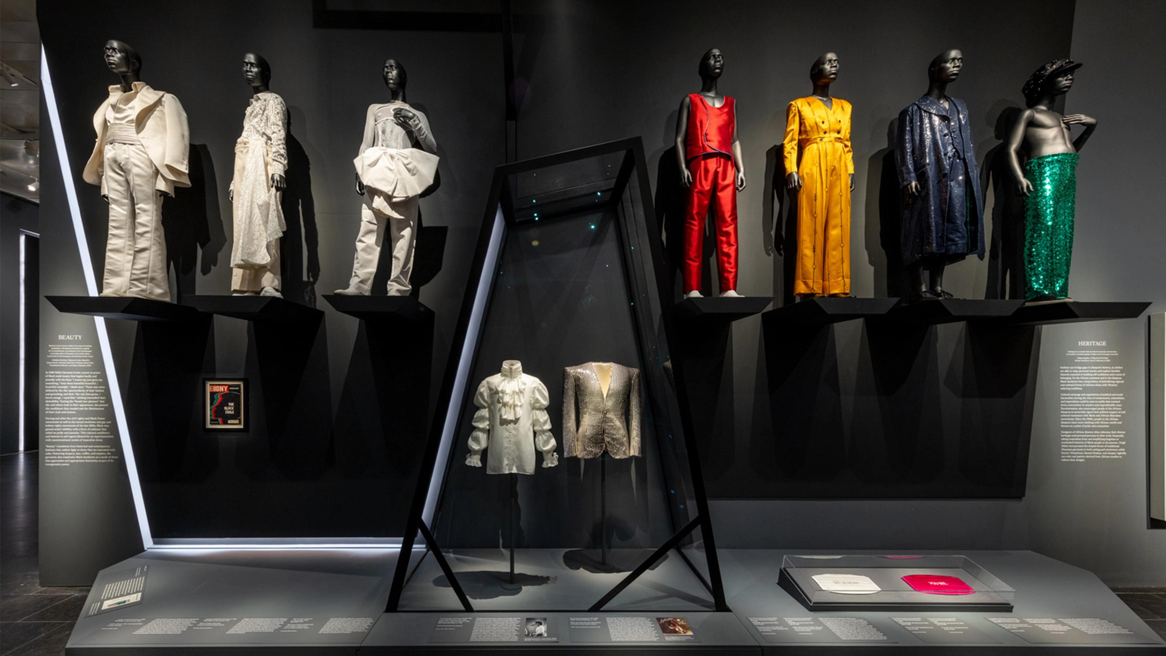 A museum exhibit displays seven mannequins in diverse, vibrant outfits, from elegant whites to vivid colors. Two detailed garments are showcased in a glass case below.