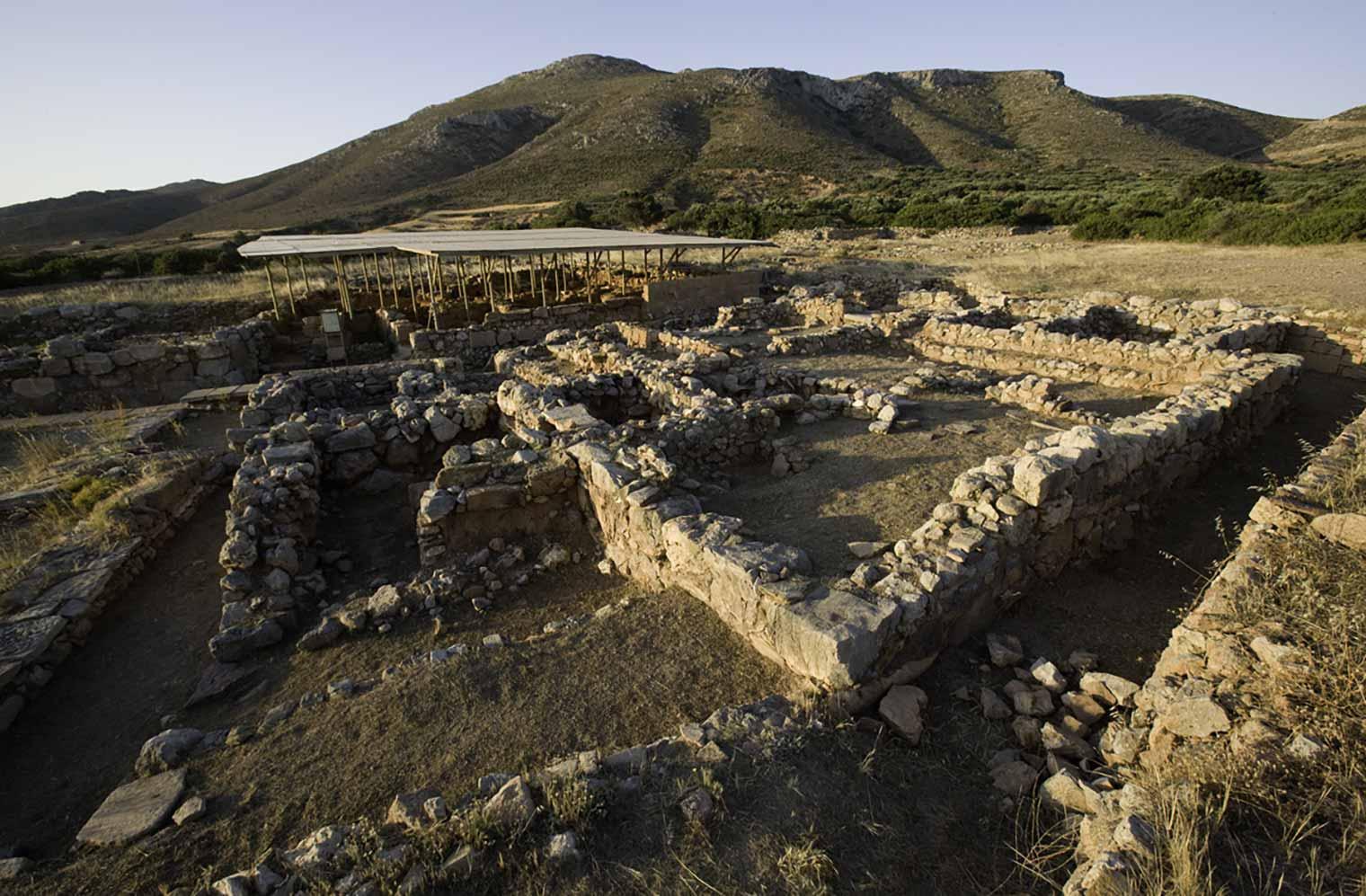 An archaeological site with stone wall foundations arranged in rectangular patterns, set in a dry, open landscape with mountains in the background and a shaded structure covering part of the ruins.