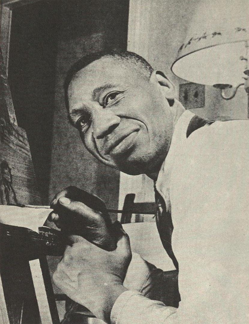 Photograph of the African American artist Horace Pippin Painting in his living room. In the photo you can see Pippin holding up his right wrist with his left hand in front of a canvas while looking at the camera. Pippin is a black man with short black hair wearing a dark vest, a tight, and a dress shirt.