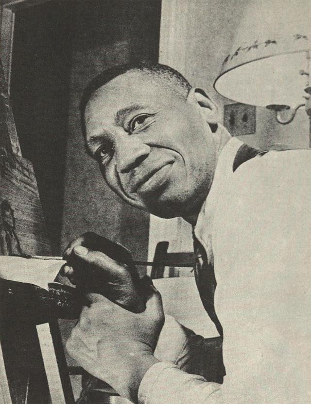 Photograph of the African American artist Horace Pippin Painting in his living room. In the photo you can see Pippin holding up his right wrist with his left hand in front of a canvas while looking at the camera. Pippin is a black man with short black hair wearing a dark vest, a tight, and a dress shirt.