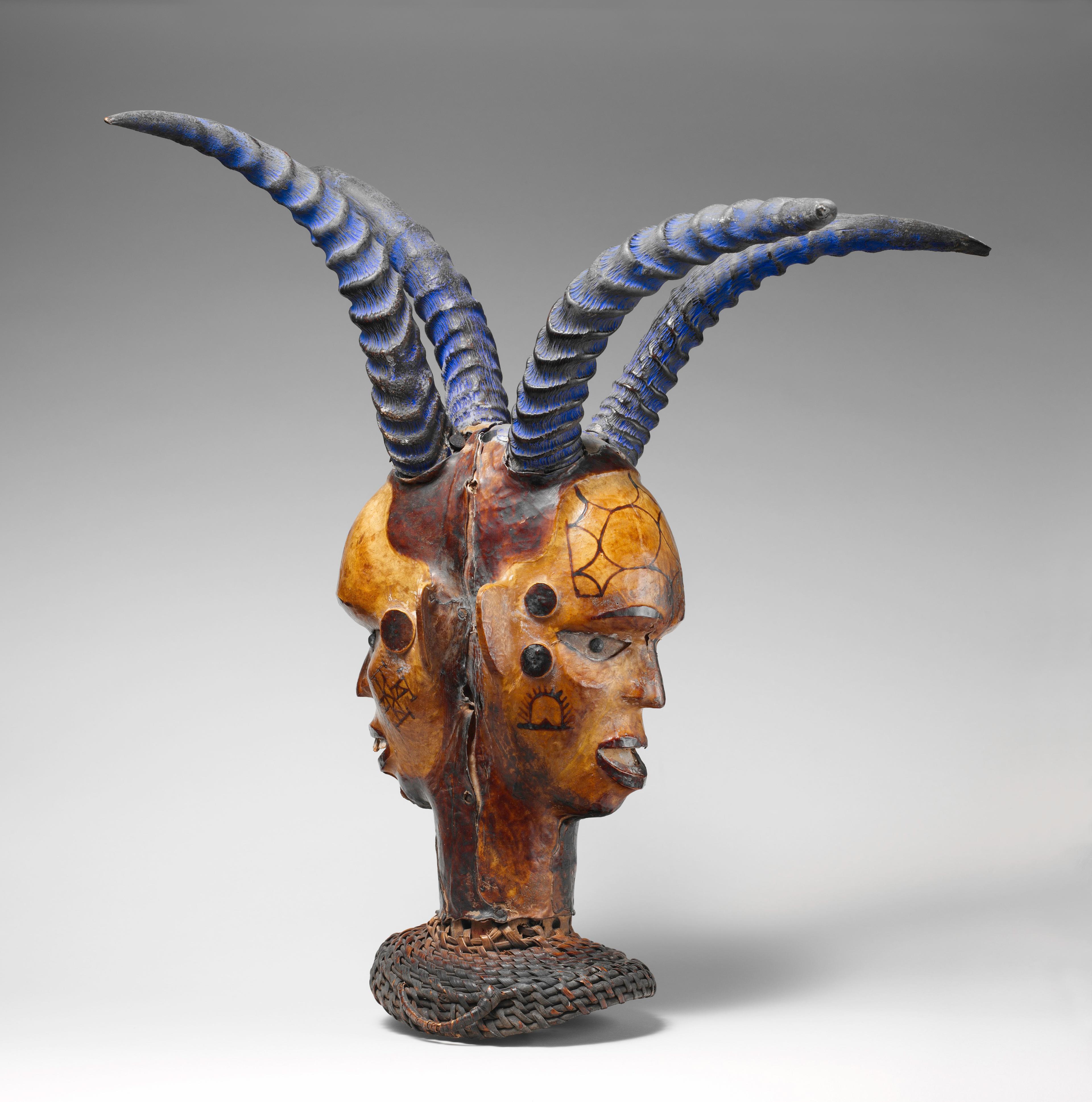 A two-faced dance crest made from untanned animal skin, with two human-like faces shown in profile and four arching blue horns.