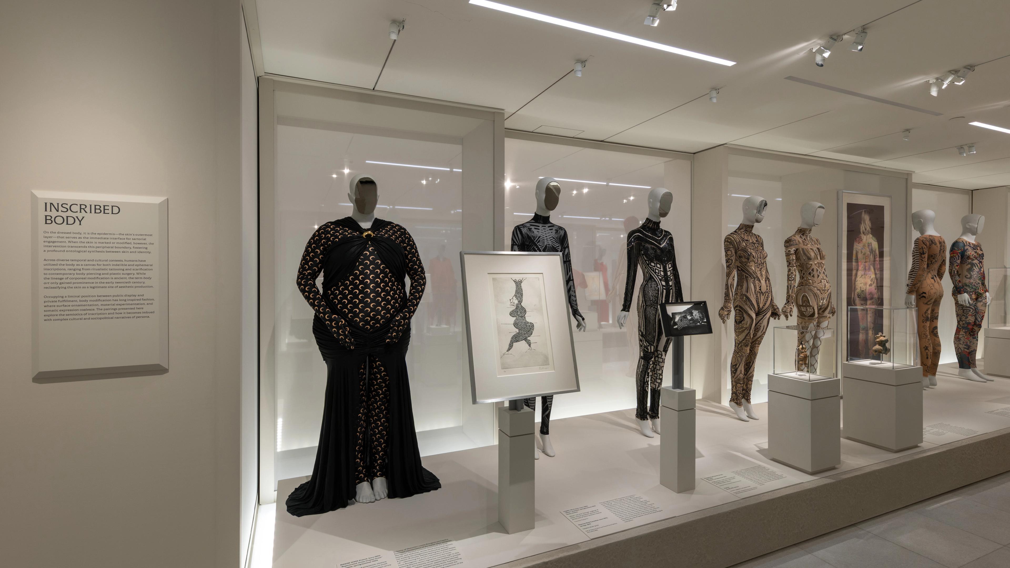 A museum exhibit displays mannequins in patterned bodysuits, with framed art on pedestals