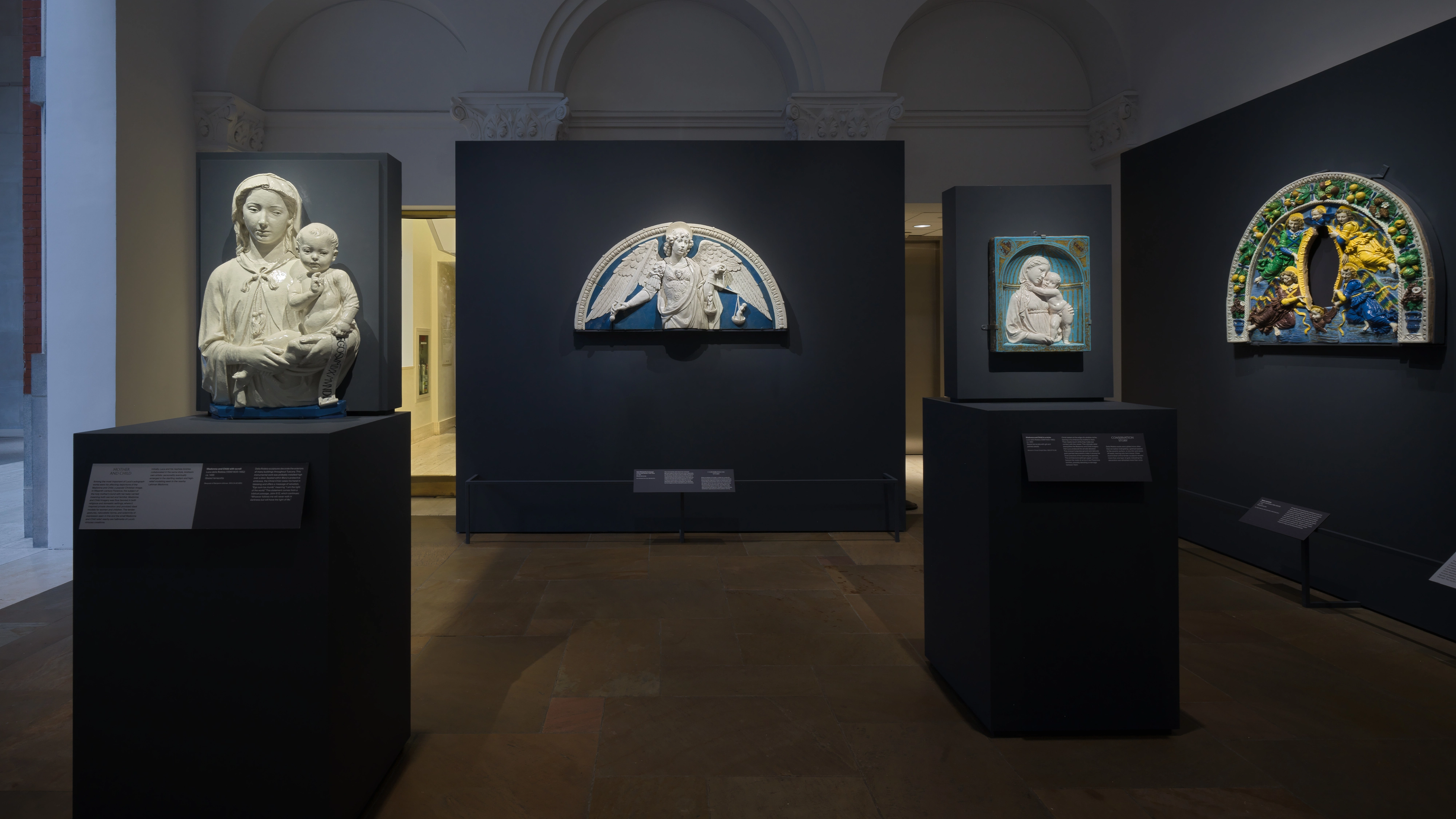 A dim museum gallery displays four religious sculptures on black pedestals, including Madonna and Child reliefs.