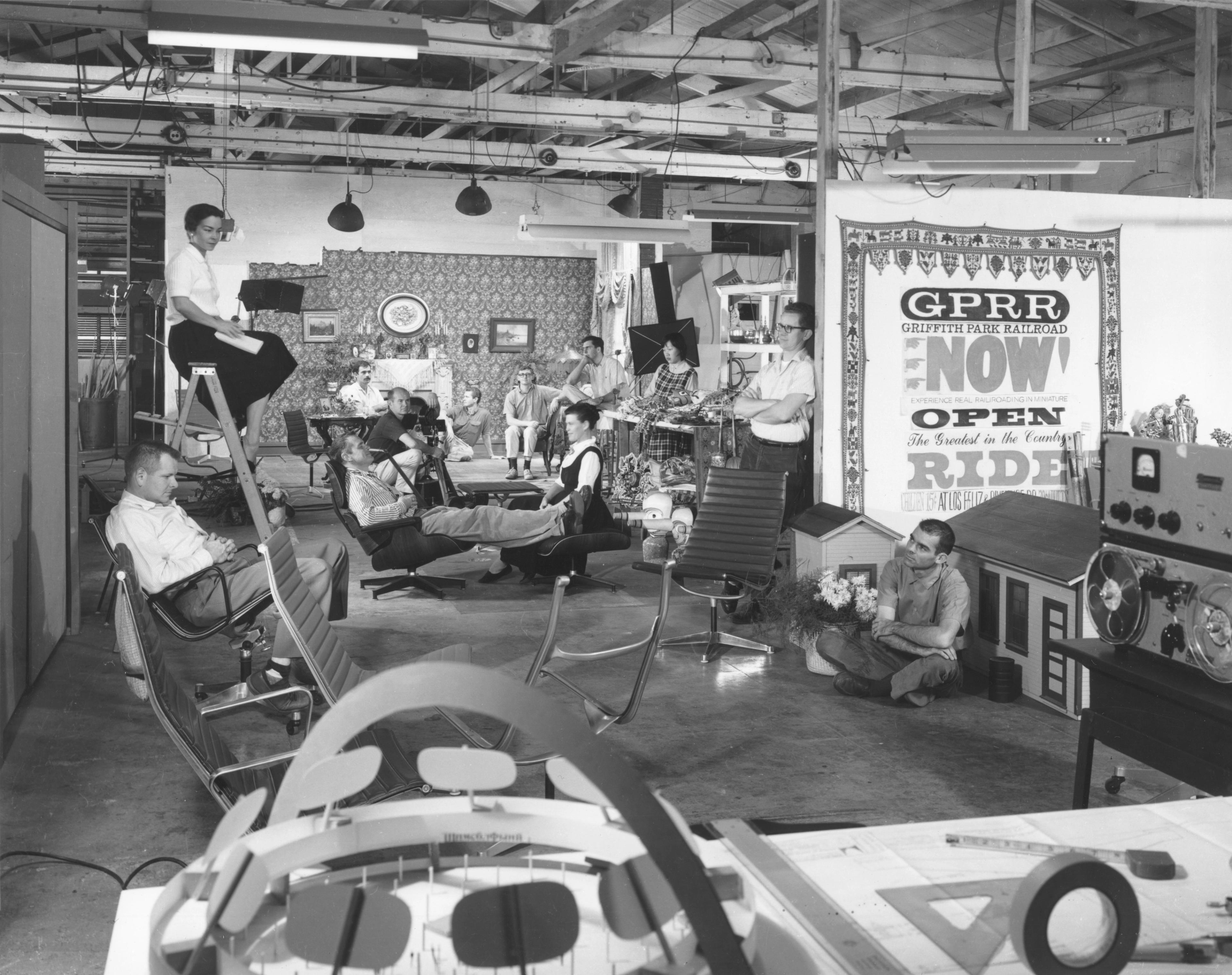Lively photograph of the inside of the Eames office, with wood-beamed ceilings and a large team of employees sitting in different Eames chairs