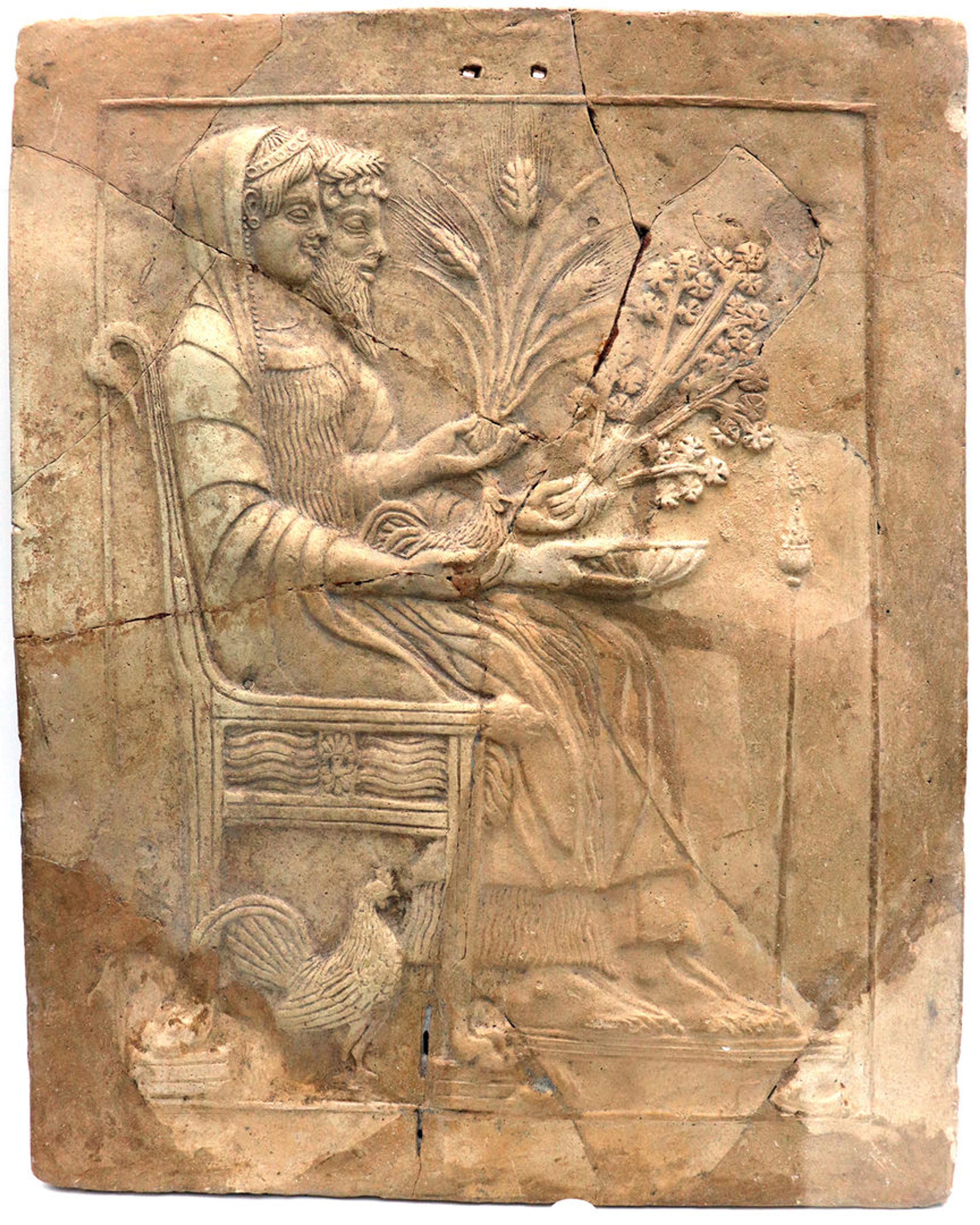 A stone votive plaque that shows Hades and Persephone seated on a throne. They hold a flat bowl called a phiale, agricultural crops, and a rooster. An additional rooster sits next to the throne.