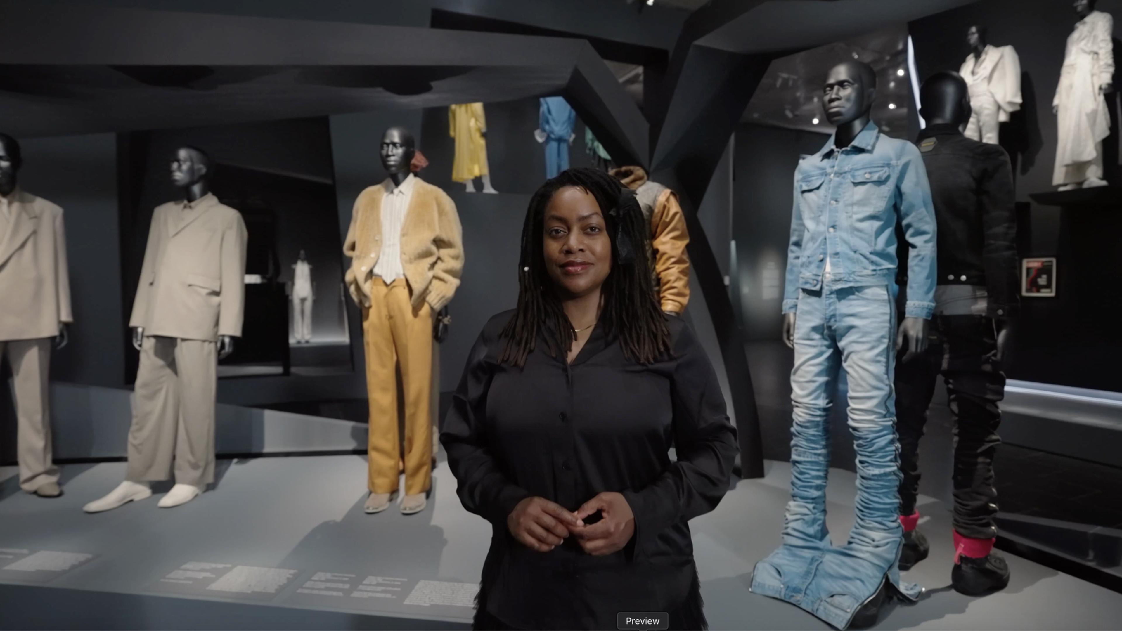Black woman wearing all black, standing in front of mannequins dressed in blue, yellow and beige.