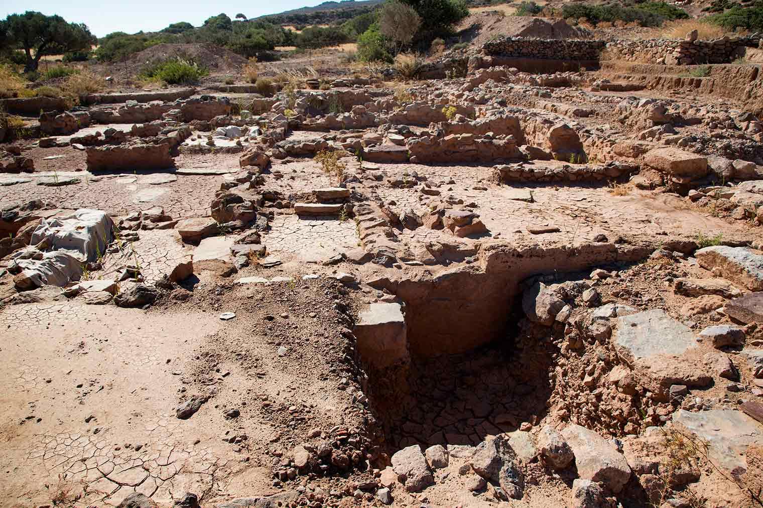 An archaeological excavation site featuring partially unearthed stone walls, dry cracked soil, and remnants of ancient structures in a sunlit, rural landscape.