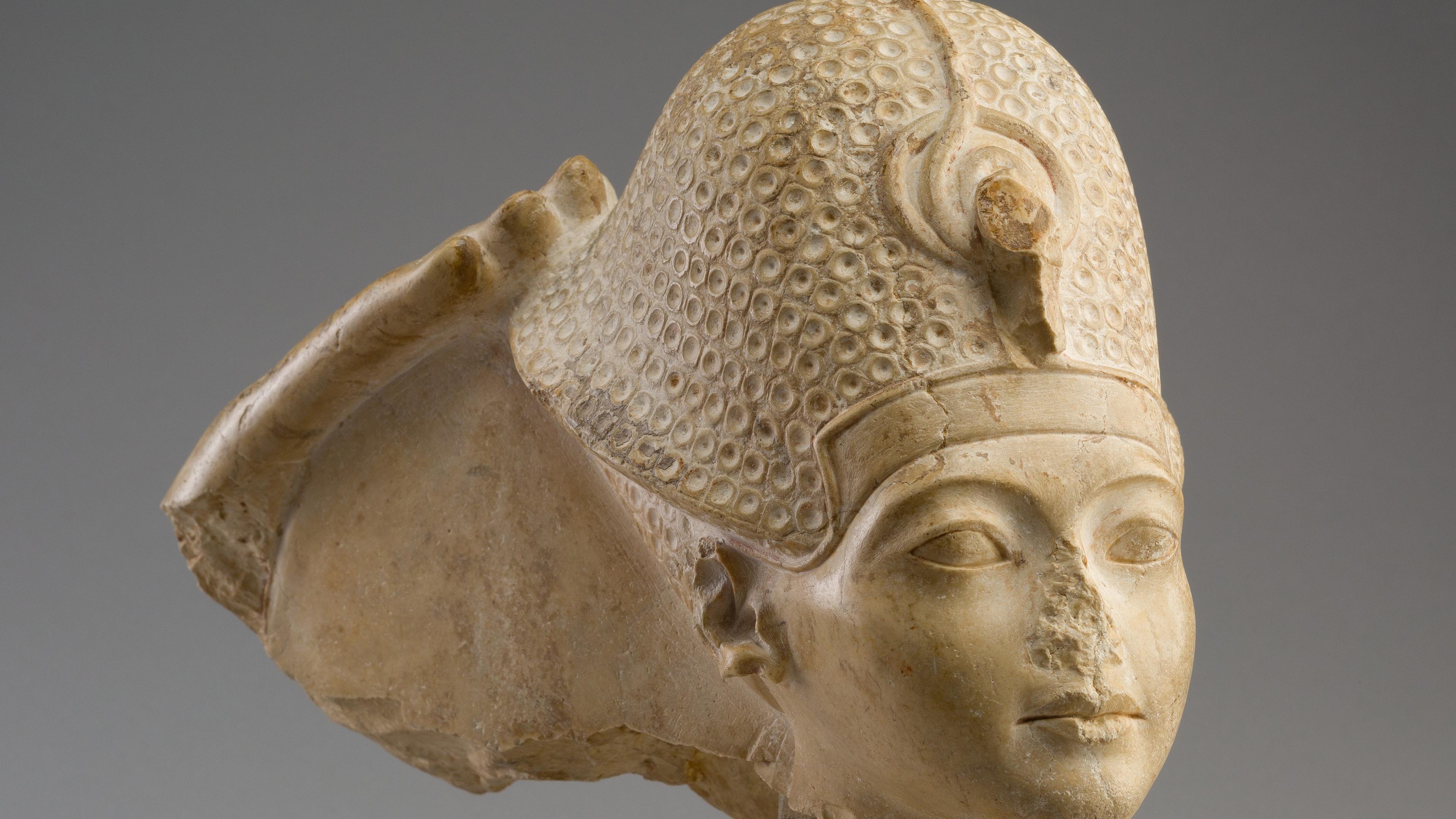 A close-up of a limestone sculpted head of Tutankhamun with young facial features and a textured and ornate helmet, set against a gray background.