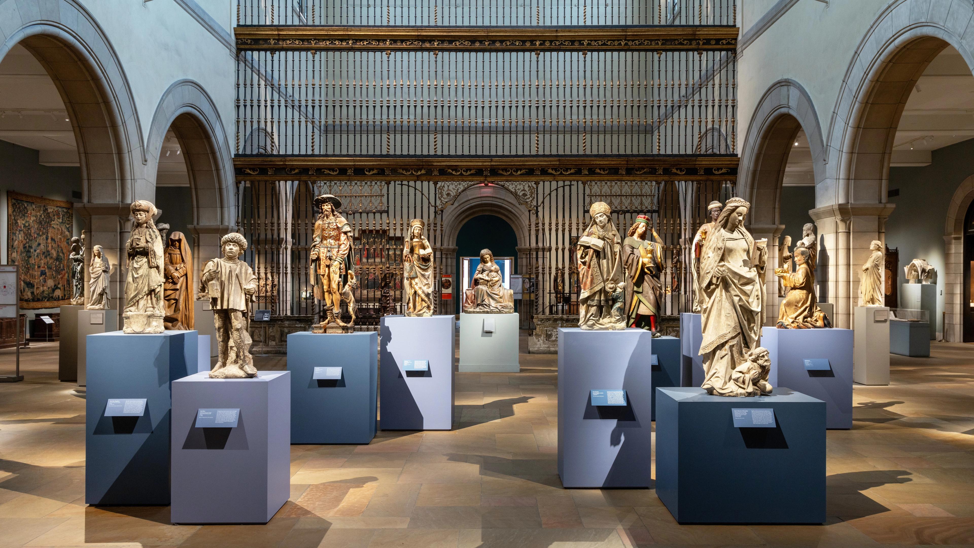 Medieval sculptures displayed on pedestals in a softly lit gallery space with stone arches and a Spanish choir screen.