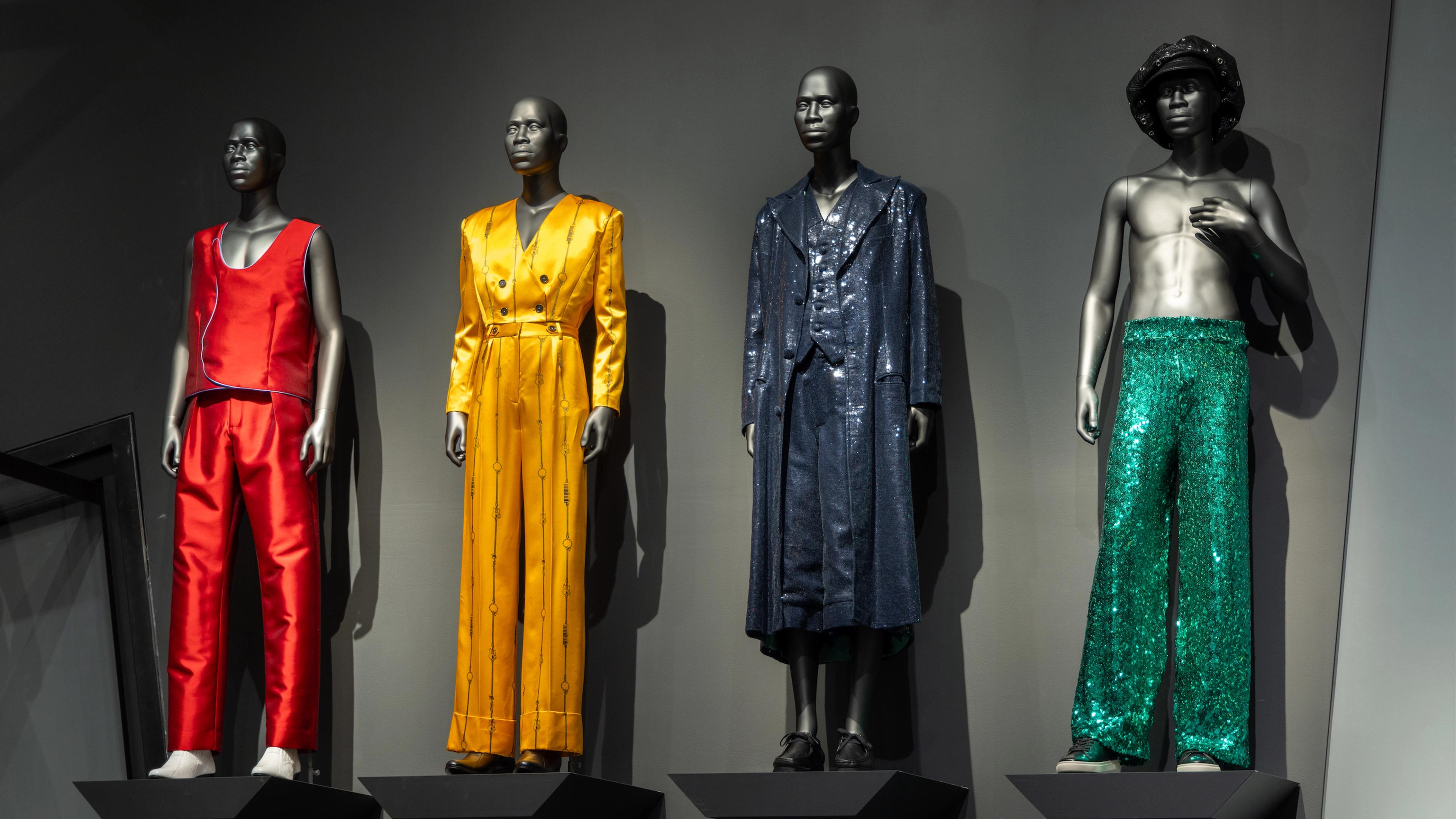 Four mannequins display vibrant fashion in a showroom. Outfits include red satin, yellow silk, blue sequins, and green sequined pants, creating a bold and stylish atmosphere.