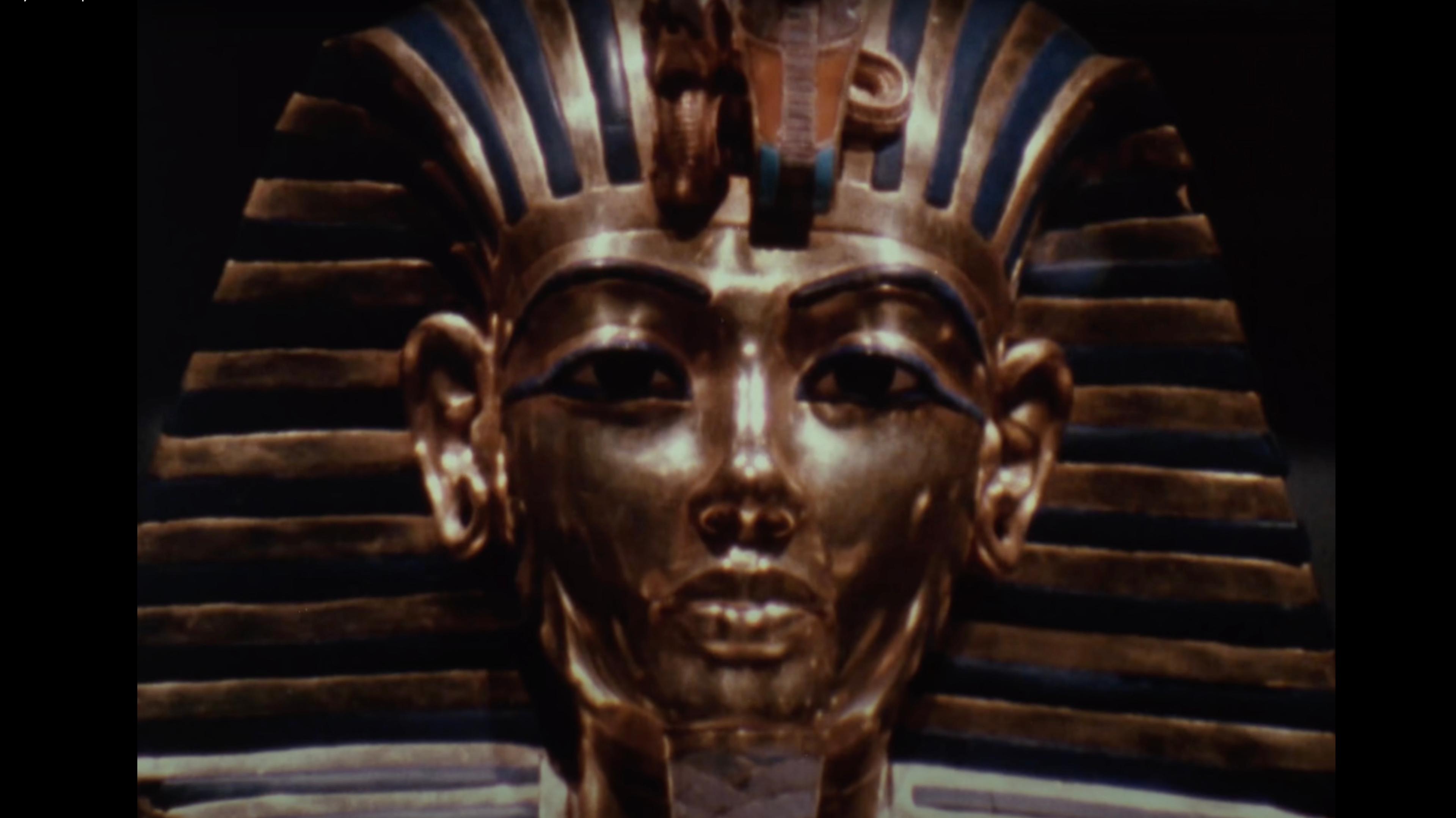 A video still of the golden funerary mask of Pharaoh Tutankhamun, adorned with blue and gold stripes and intricate detailing.