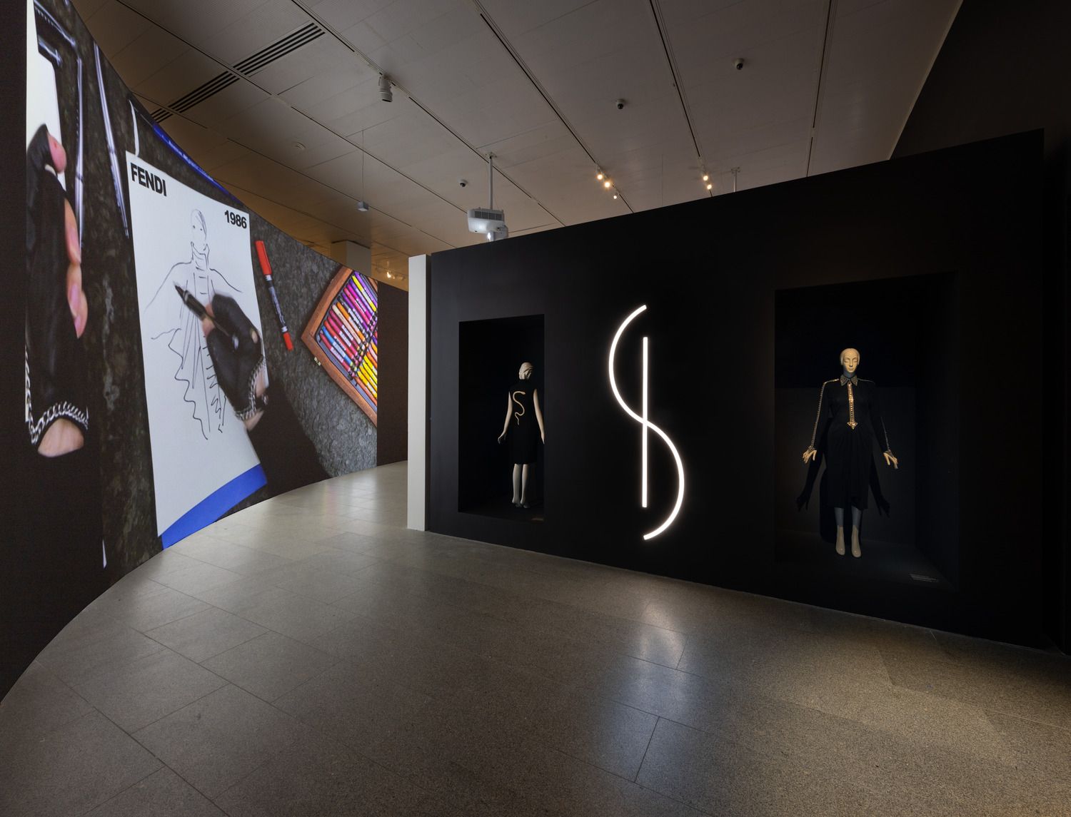 A sweeping view of a dark gallery featuring featuring two mannequins on the right and a projection of a gloved hand drawing a fashion sketch on the left.